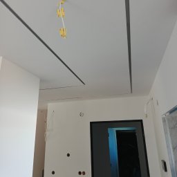 Krzysztof Wacek - Biały sufit podwieszany z szarymi listwami LED w nowym wnętrzu. Widoczne kable elektryczne i otwory w ścianach. W tle drzwi i schody.