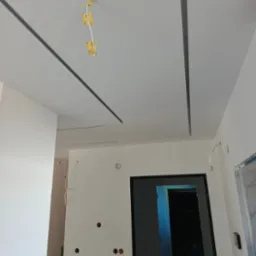 Biały sufit podwieszany z szarymi listwami LED w nowym wnętrzu. Widoczne kable elektryczne i otwory w ścianach. W tle drzwi i schody.