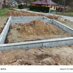 Fundamenty domu w trakcie budowy, wykonane z szarych bloczków betonowych, na tle ziemi i zieleni. Widoczny stos ziemi wewnątrz fundamentów. Budowa domu w toku.