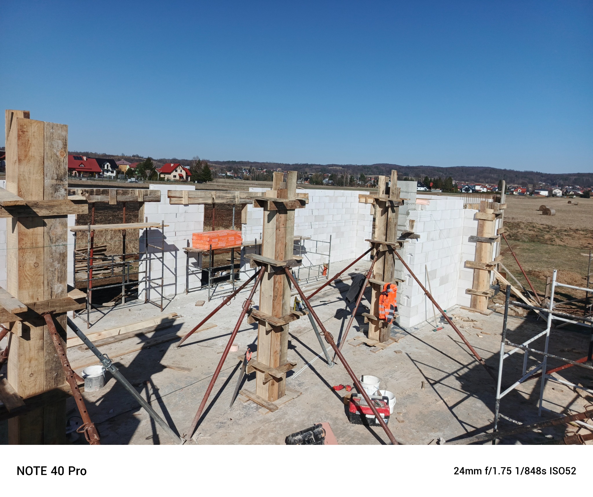 Wznoszenie ścian z białych bloczków, widoczne drewniane stemple podtrzymujące konstrukcję stropu, pomarańczowa skrzynka i kamizelka odblaskowa, rusztowanie, w tle zabudowa jednorodzinna i pola.