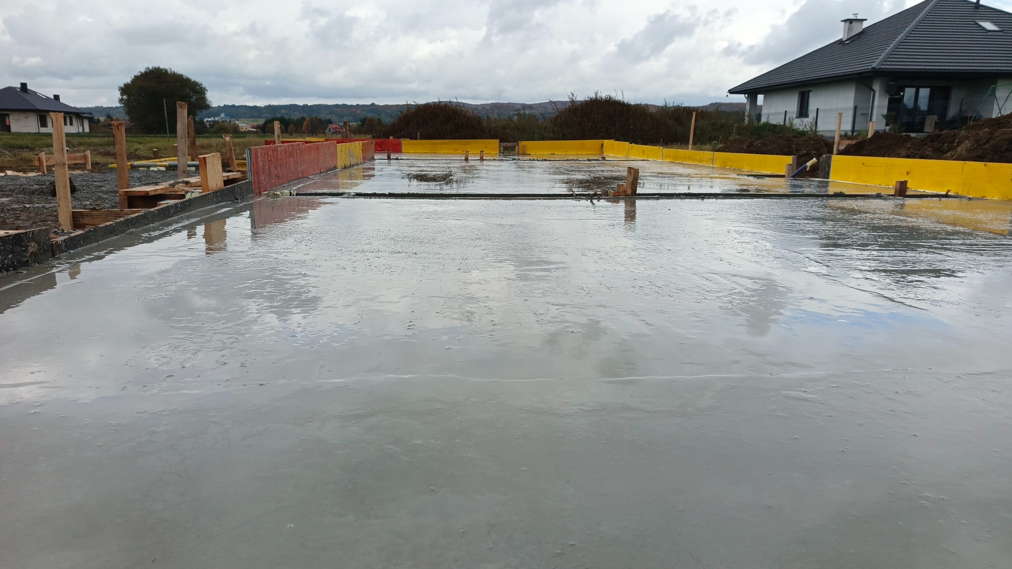 Świeżo wylany fundament domu jednorodzinnego z widocznym szalunkiem w kolorze żółtym i czerwonym, odbijającym pochmurne niebo. W tle dom w budowie i zieleń.