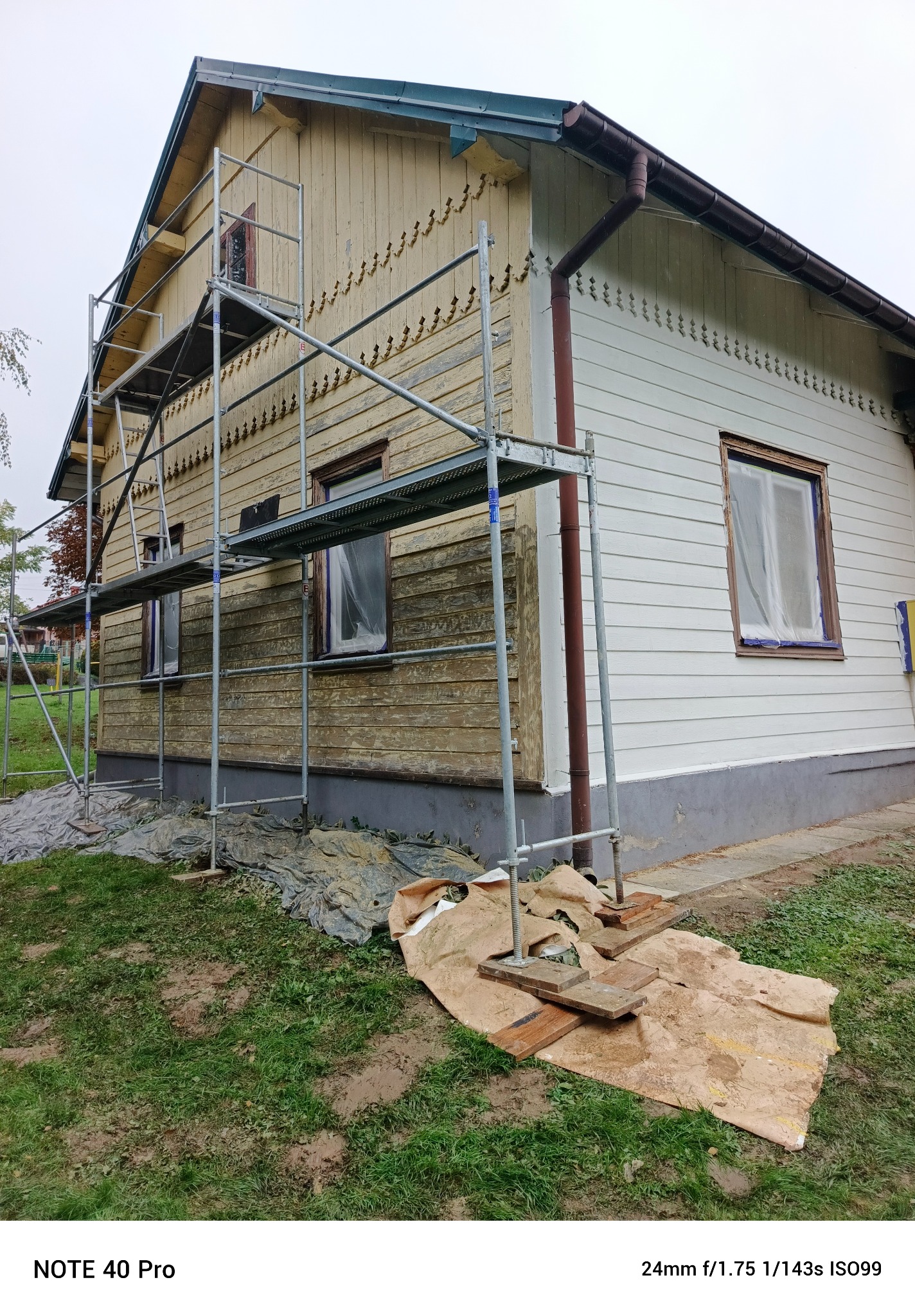 Renowacja drewnianego domu z dekoracyjnymi elementami. Część elewacji pomalowana na biało, druga w trakcie przygotowania. Rusztowanie, zabezpieczone okna.