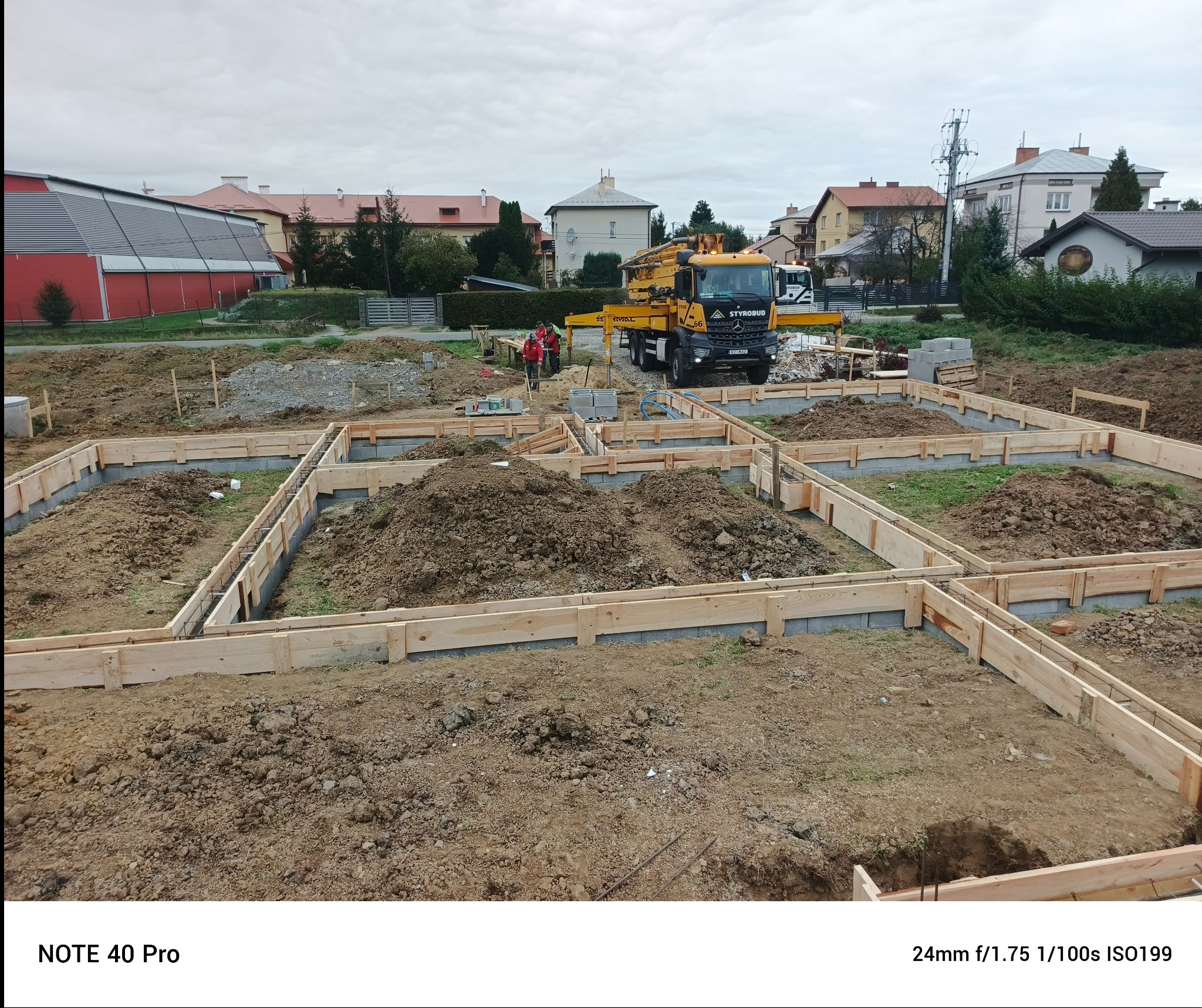 Przygotowanie fundamentów pod budowę domu: drewniane szalunki, zbrojenie i betonowa pompa Styrobud w tle. Widoczni pracownicy w czerwonych kurtkach.