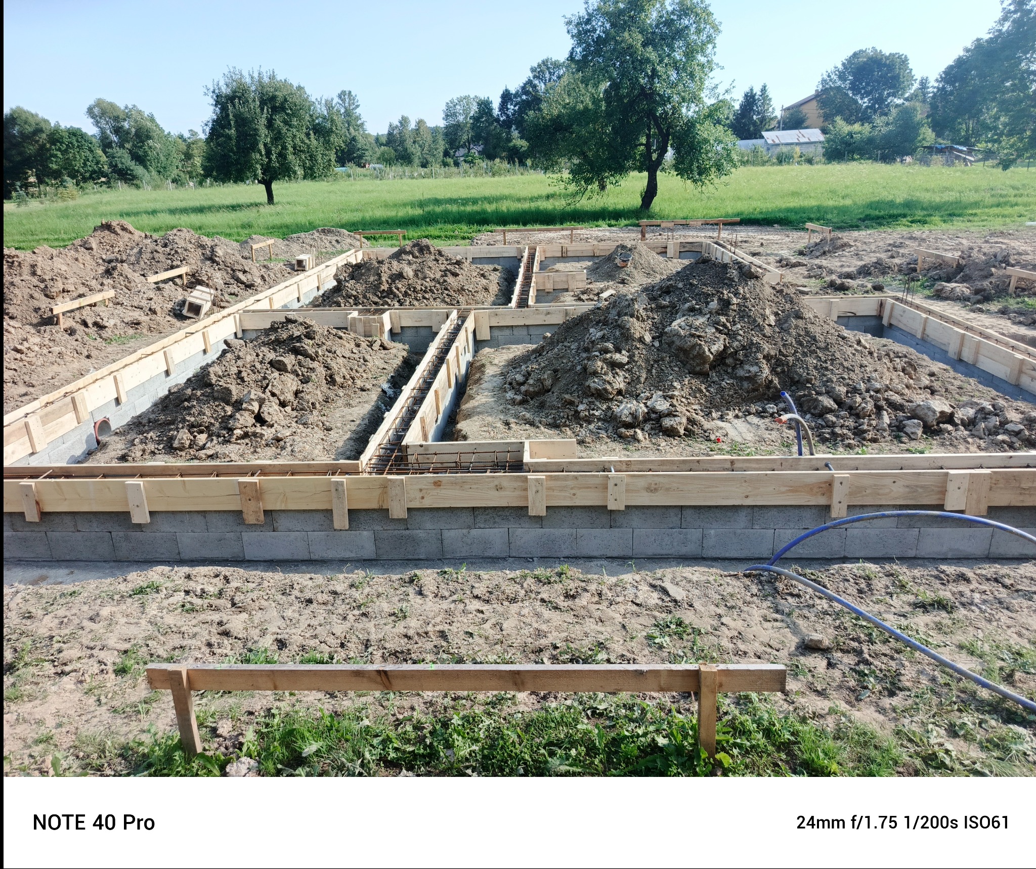 Ziemia w wykopach fundamentowych zbrojonych stalą, otoczonych drewnianym szalunkiem na bloczkach betonowych, na tle zielonej łąki i drzew w słoneczny dzień.