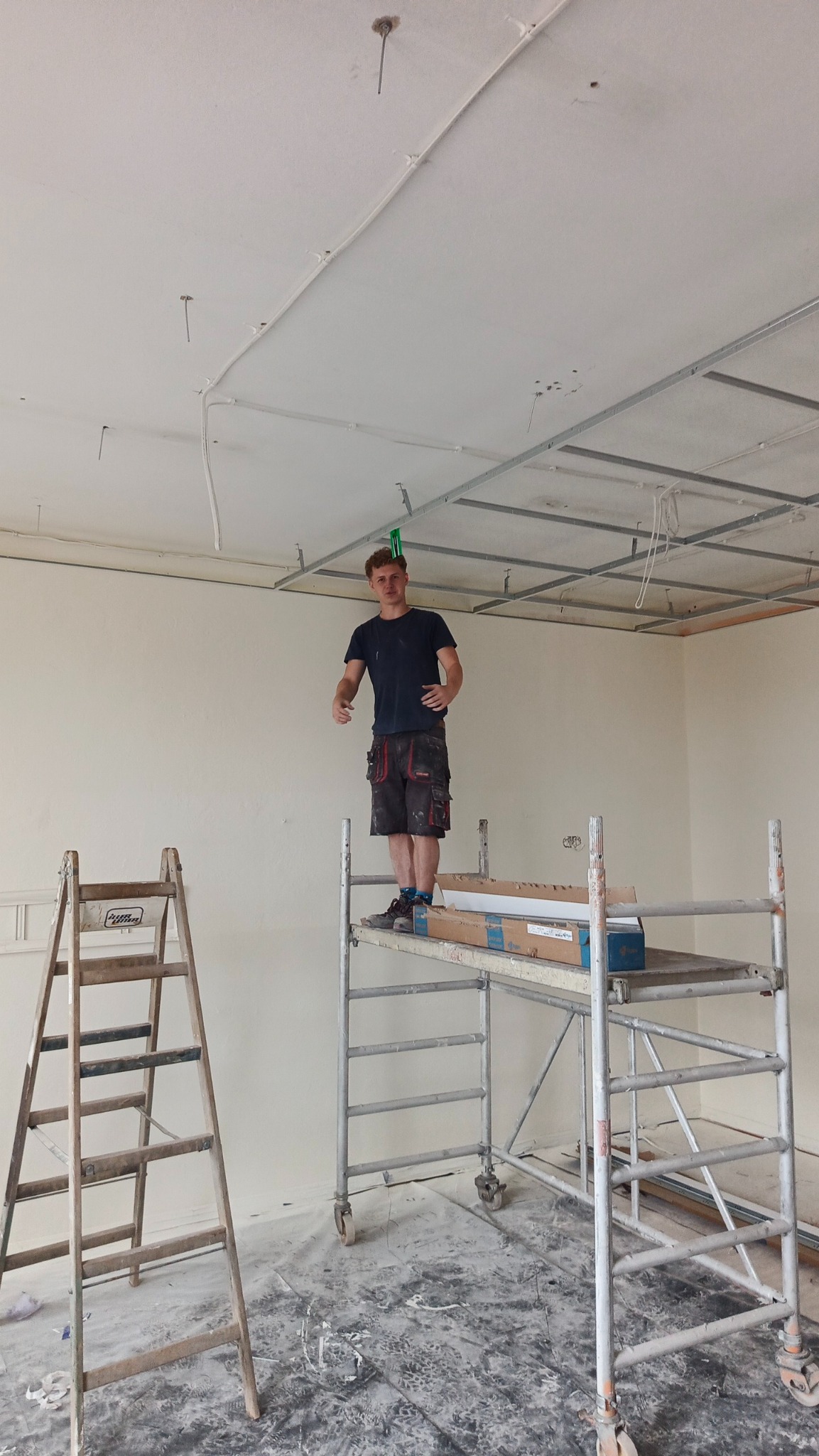 Pracownik na rusztowaniu montuje metalowy stelaż sufitu podwieszanego w jasnym wnętrzu. Widoczna drabina, kable i częściowo zamontowana płyta.