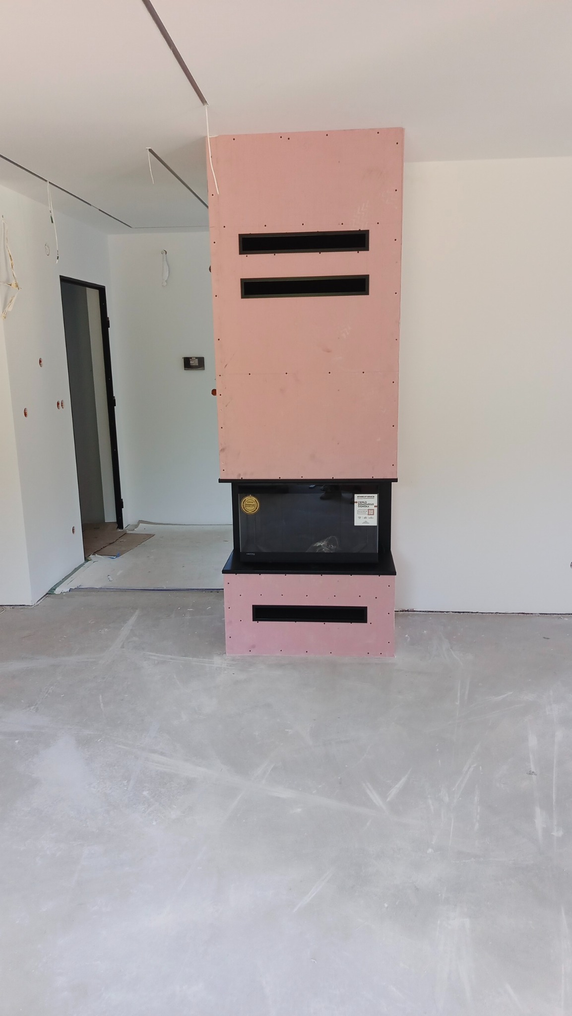 Wnętrze z obudową kominka z płyty karton-gips w kolorze różowym, z czarnymi wnękami wentylacyjnymi. Podłoga szara, surowa. Widoczne niedokończone prace wykończeniowe.