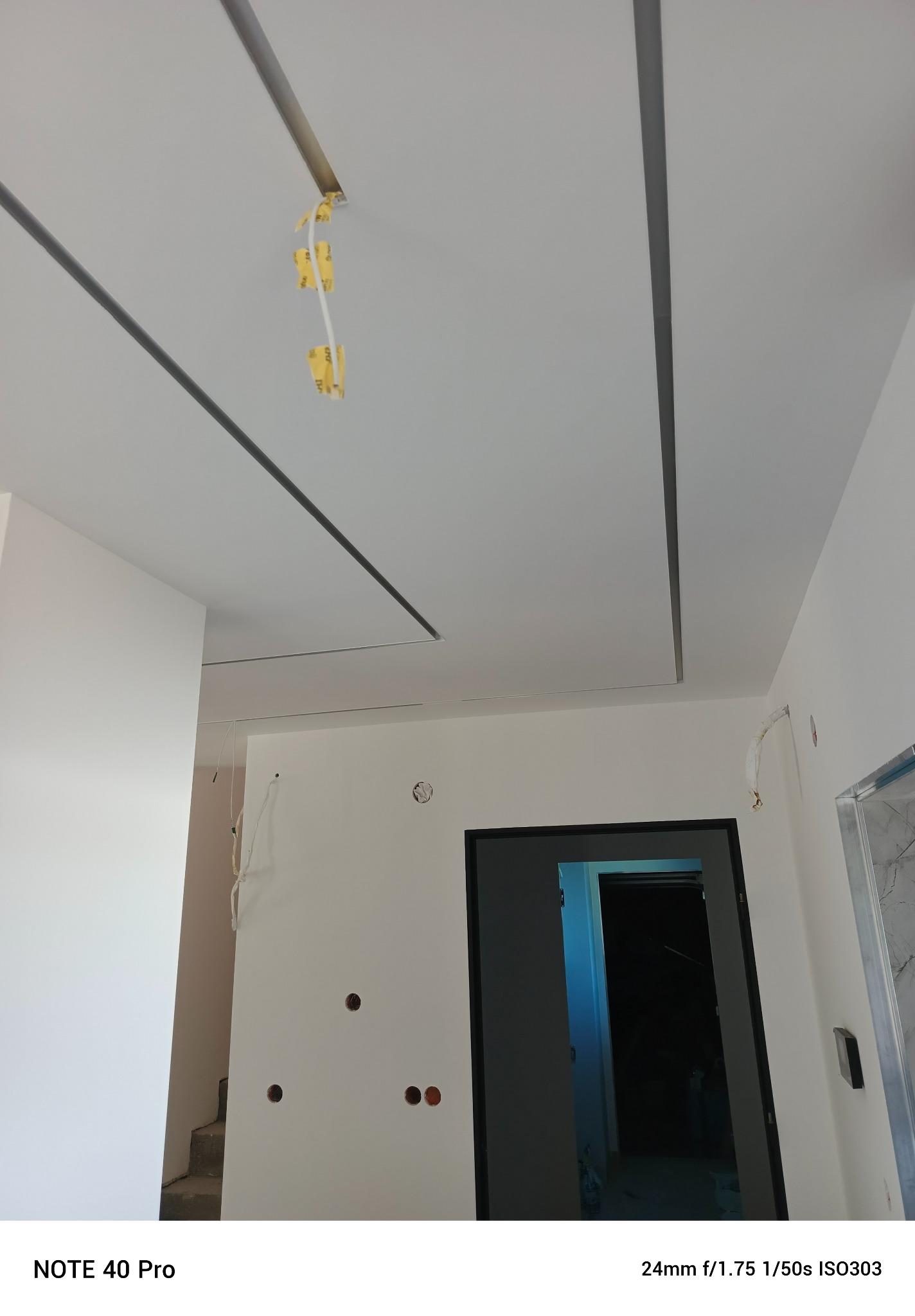 Biały sufit podwieszany z szarymi listwami LED w nowym wnętrzu. Widoczne kable elektryczne i otwory w ścianach. W tle drzwi i schody.