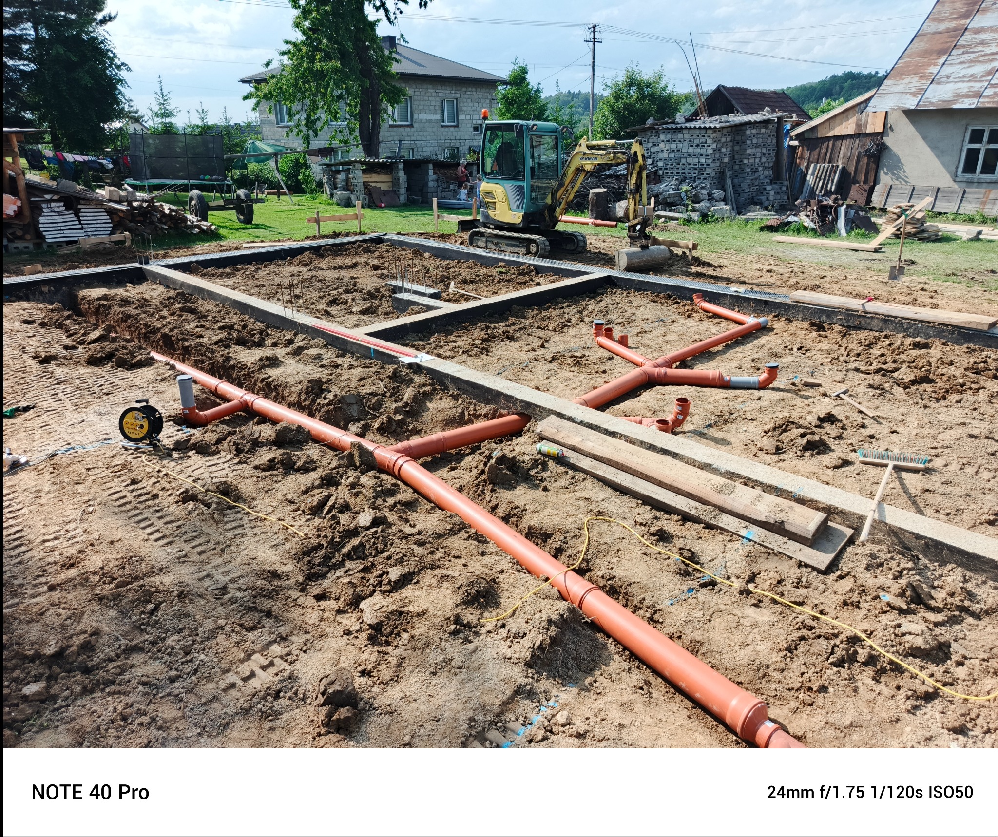 Fundamenty domu w trakcie budowy: widoczne rury kanalizacyjne, szalunki, ziemia i mini koparka w tle. Prace ziemne i instalacyjne w toku.