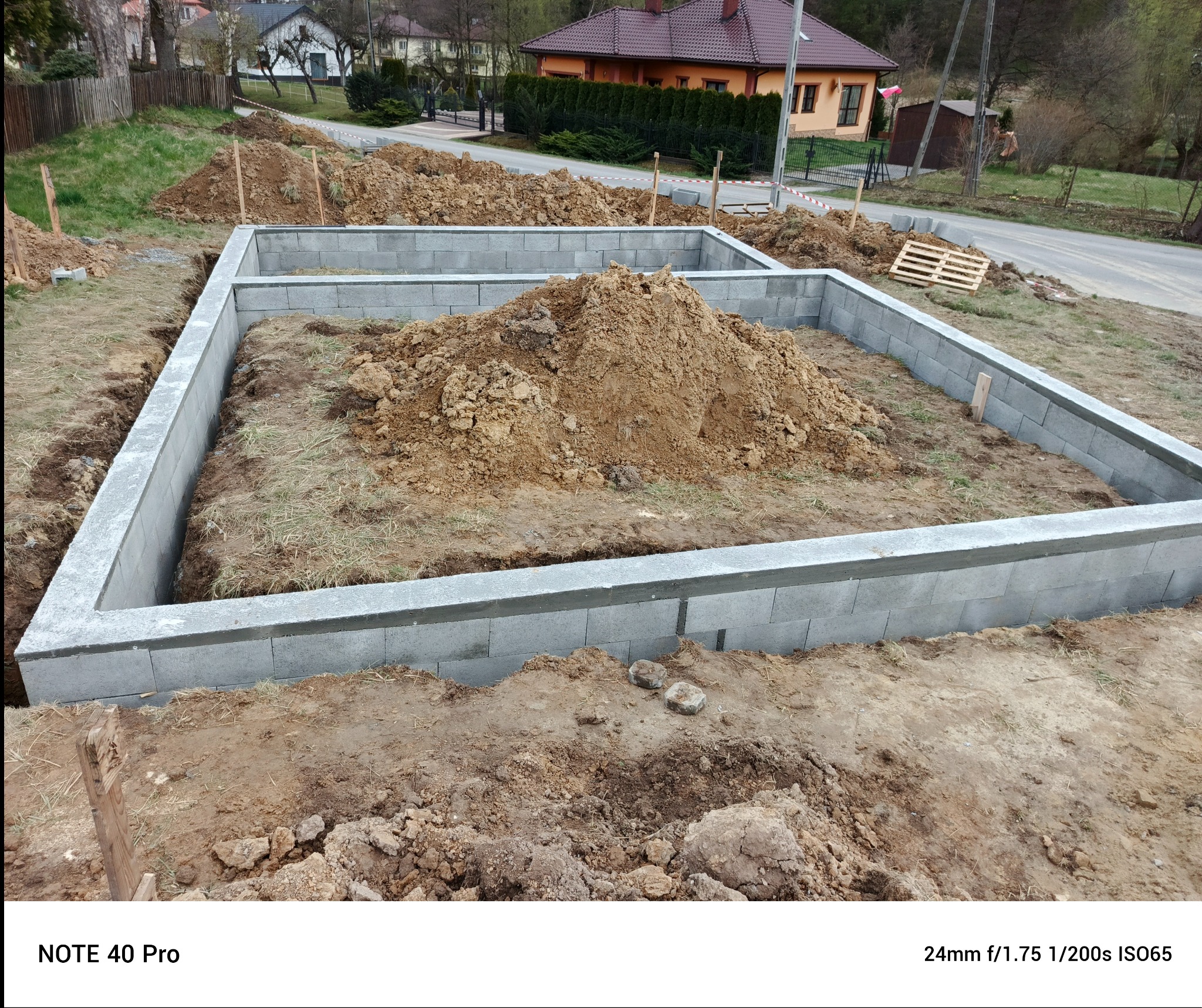 Fundamenty domu w trakcie budowy, wykonane z szarych bloczków betonowych, na tle ziemi i zieleni. Widoczny stos ziemi wewnątrz fundamentów. Budowa domu w toku.