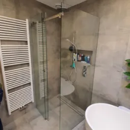 Nowoczesna łazienka z kabiną prysznicową typu walk-in, grzejnikiem drabinkowym i wolnostojącą wanną. Ściany wykończone płytami imitującymi beton.