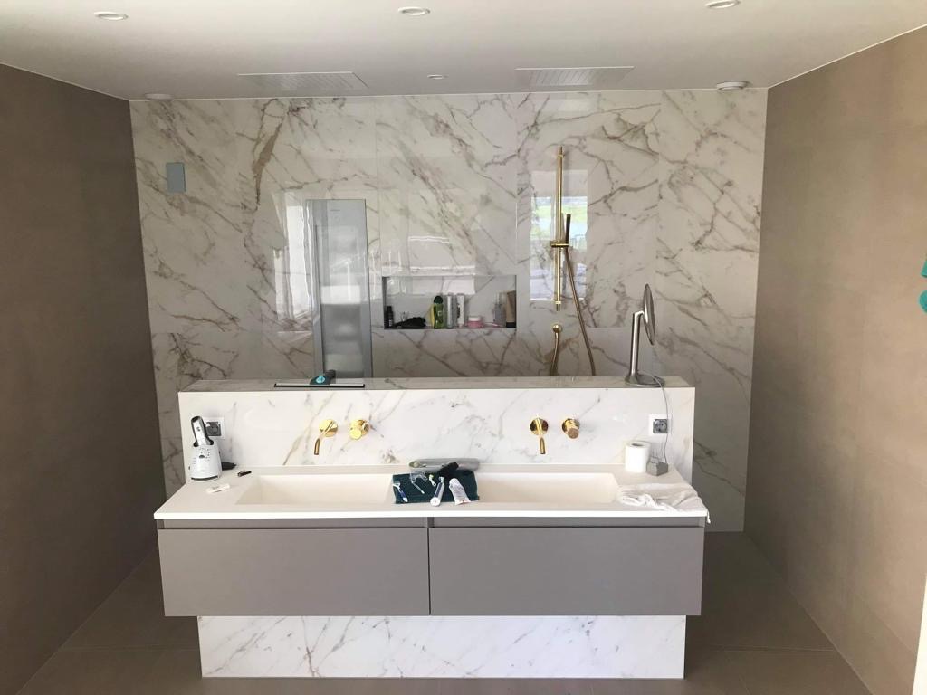 Elegancka łazienka z marmurową ścianą i złotą armaturą. Podwójna umywalka z szarą szafką. Widoczny prysznic i wnęka na kosmetyki. Nowoczesny design.