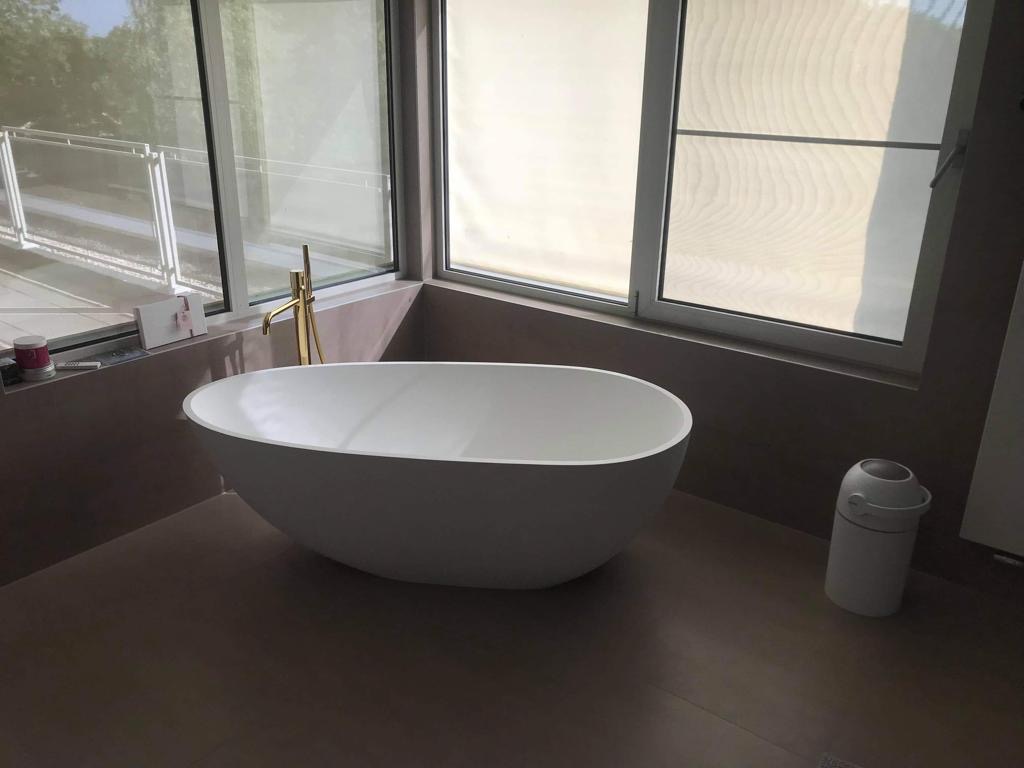 Nowoczesna łazienka z wolnostojącą białą wanną, złotą baterią i dużym oknem z widokiem na zieleń. Minimalistyczny design, brązowe płytki podłogowe i elegancki kosz na śmieci.