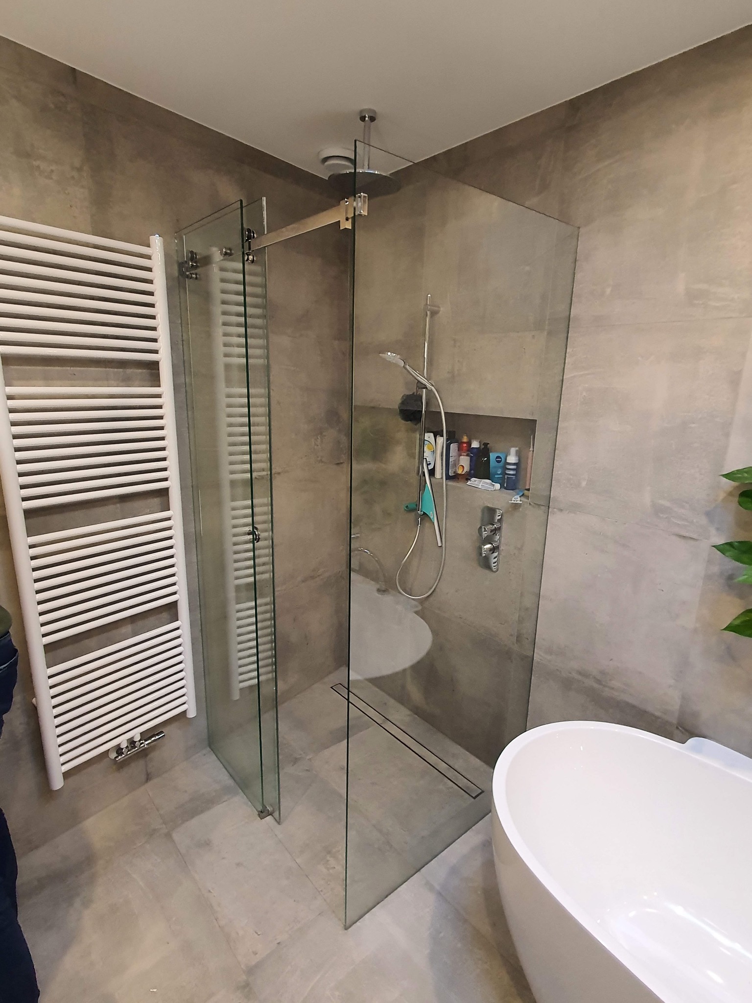 Nowoczesna łazienka z kabiną prysznicową typu walk-in, grzejnikiem drabinkowym i wolnostojącą wanną. Ściany wykończone płytami imitującymi beton.