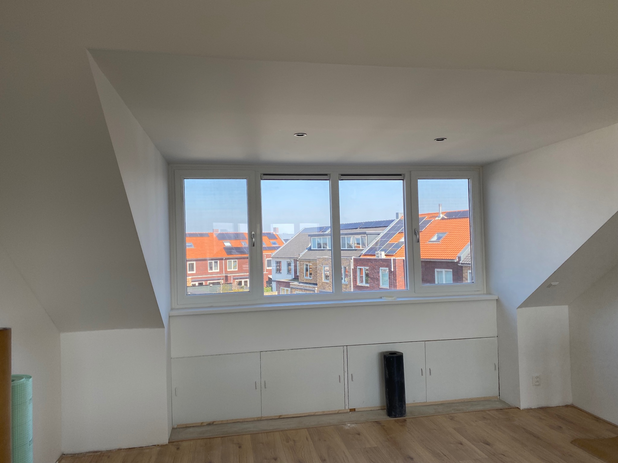 Poddasze po remoncie: białe ściany, okno z widokiem na zabudowę, zabudowa pod oknem z białymi szafkami, jasna podłoga. Widoczne rolki materiału budowlanego.