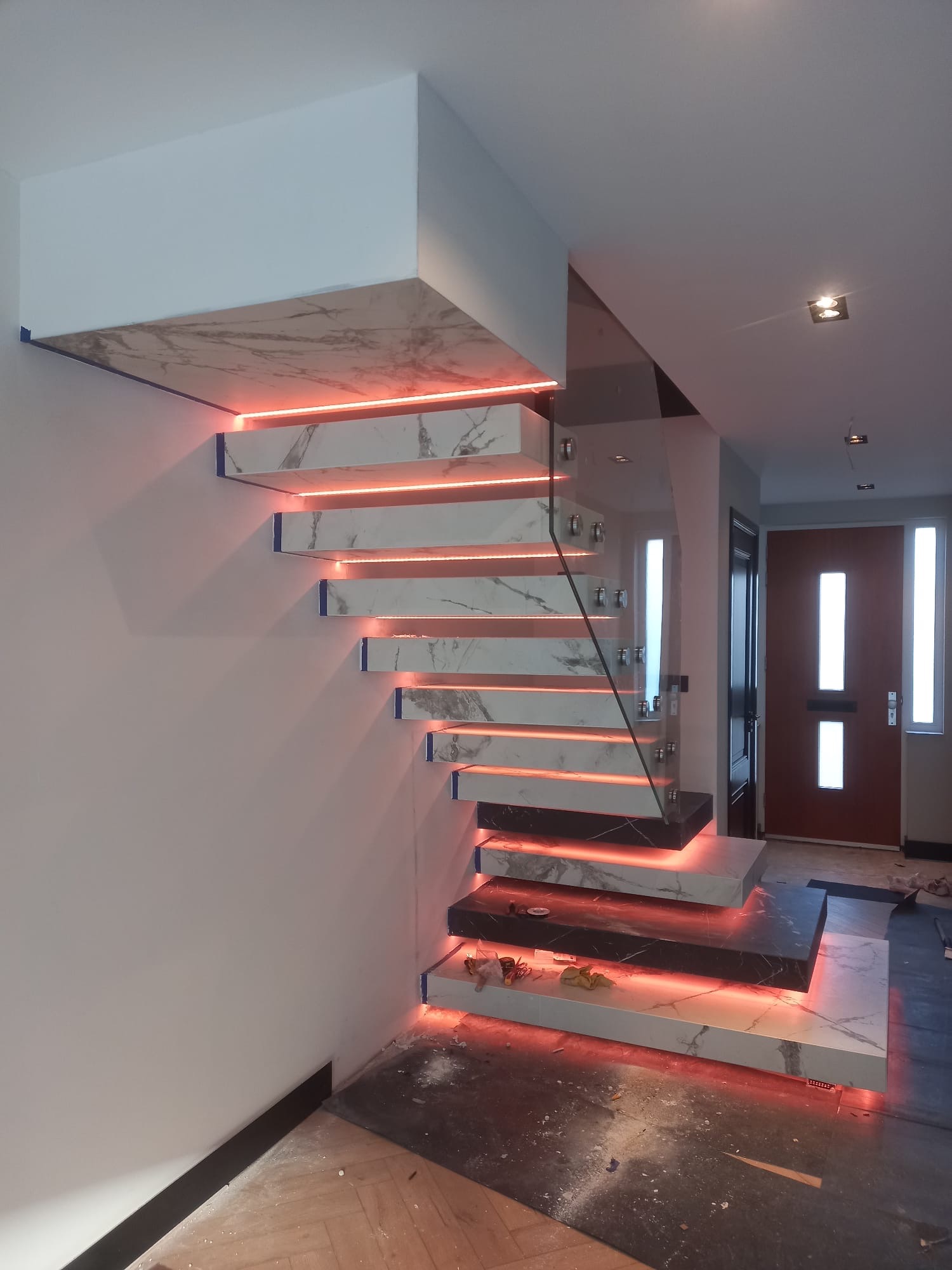 Nowoczesne, podświetlane schody z marmurowymi stopniami i szklaną balustradą w jasnym wnętrzu. Widoczne detale wykończenia i oświetlenie LED.