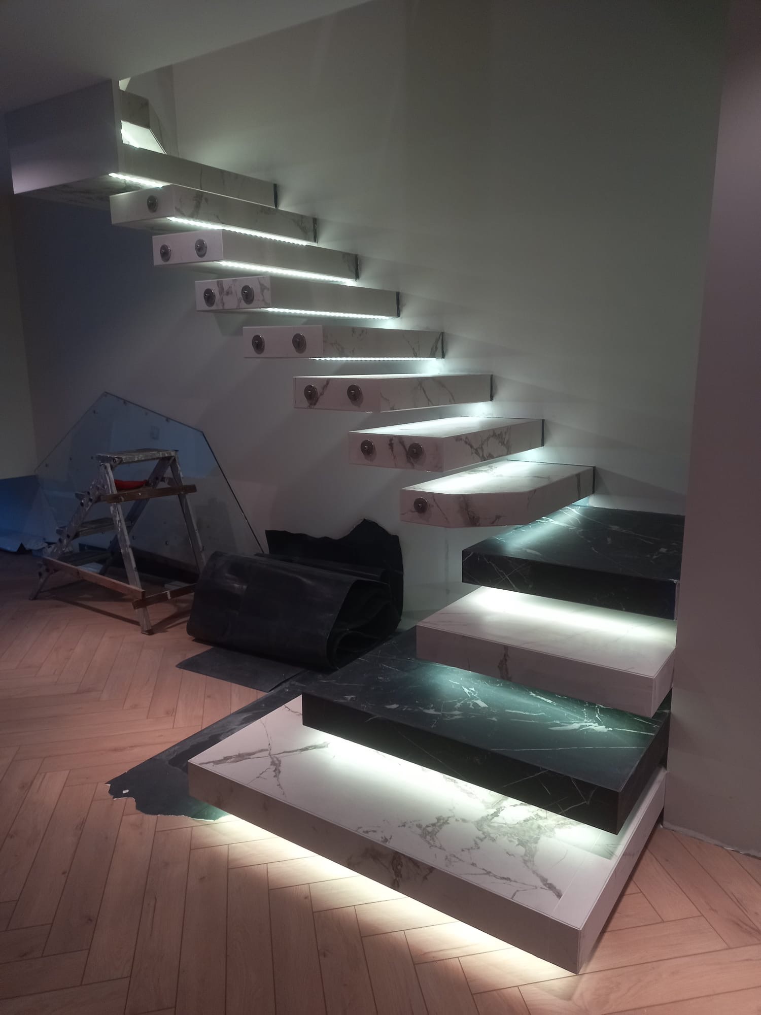 Nowoczesne, marmurowe schody wspornikowe z podświetleniem LED, widoczne z boku, z elementami wykończeniowymi i narzędziami w tle, na drewnianej podłodze.