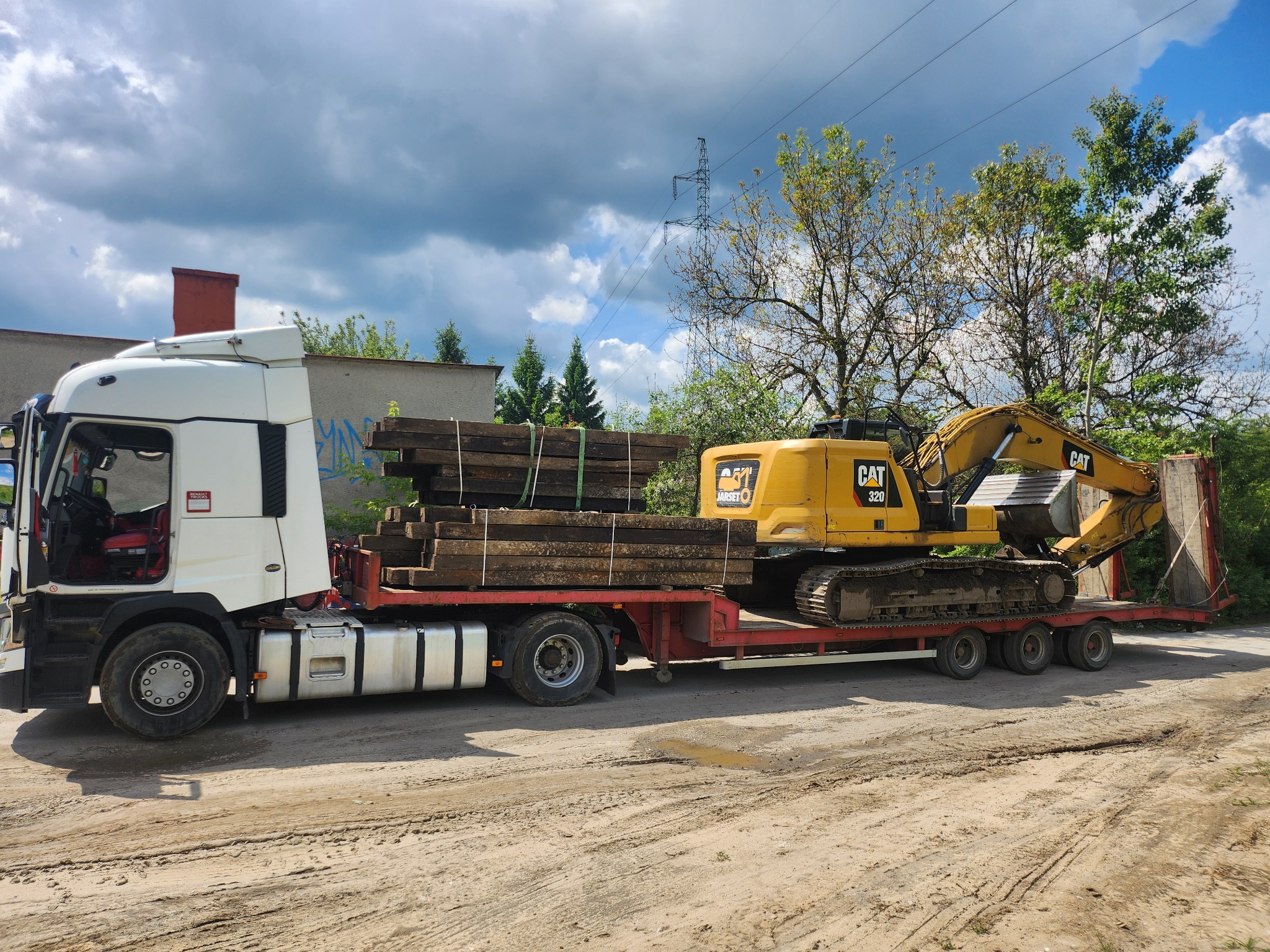 Laweta z żółtą koparką CAT 320 i drewnianymi belkami na czerwonej platformie, biała kabina ciężarówki, szare chmury w tle, teren piaszczysty.