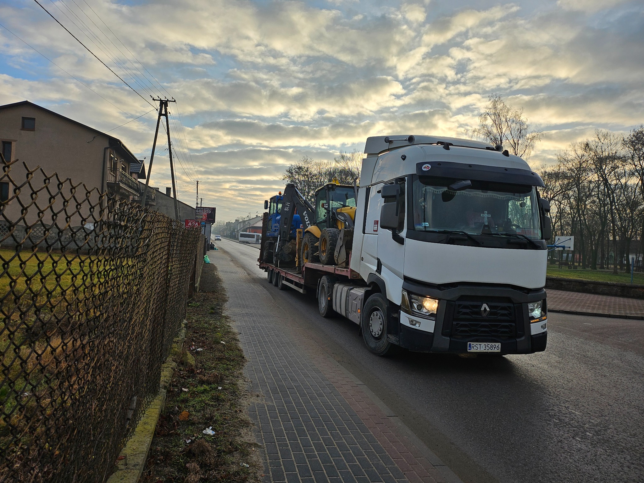 Laweta przewozi dwie maszyny budowlane (koparki) na niskopodwoziowej platformie. Widok z boku na ciężarówkę na tle pochmurnego nieba i zabudowań.