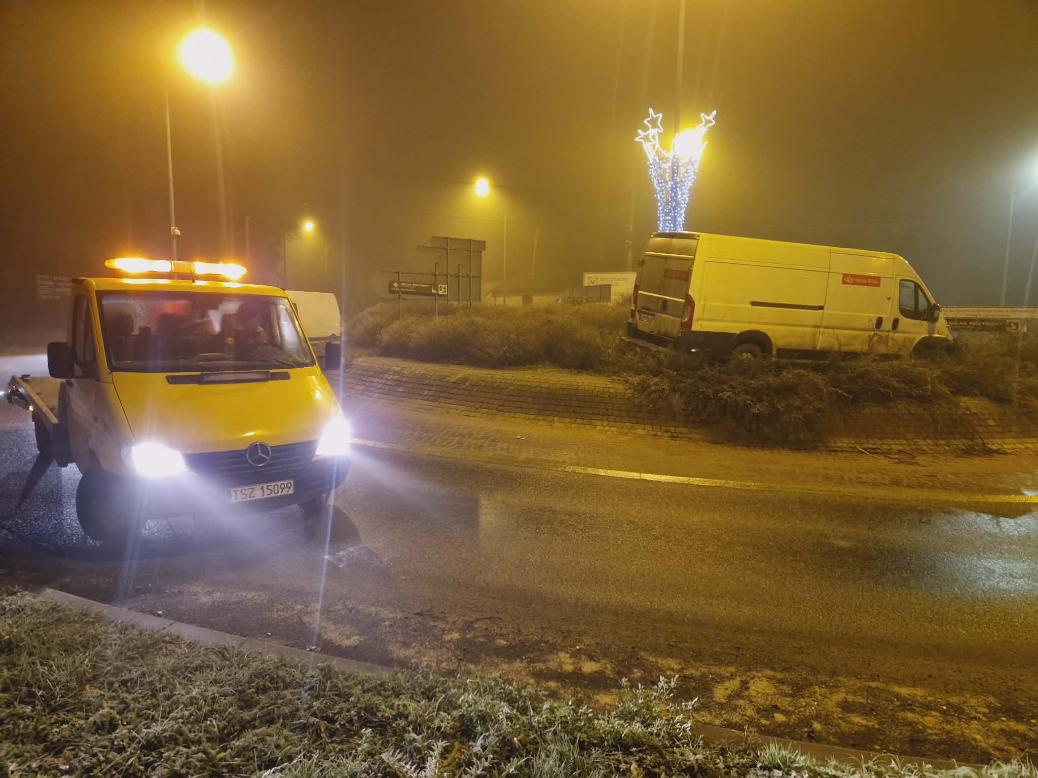 Żółta laweta Mercedes z włączonymi światłami awaryjnymi na drodze w nocy, w tle uszkodzony biały bus na rondzie z dekoracją świąteczną. Mgła.