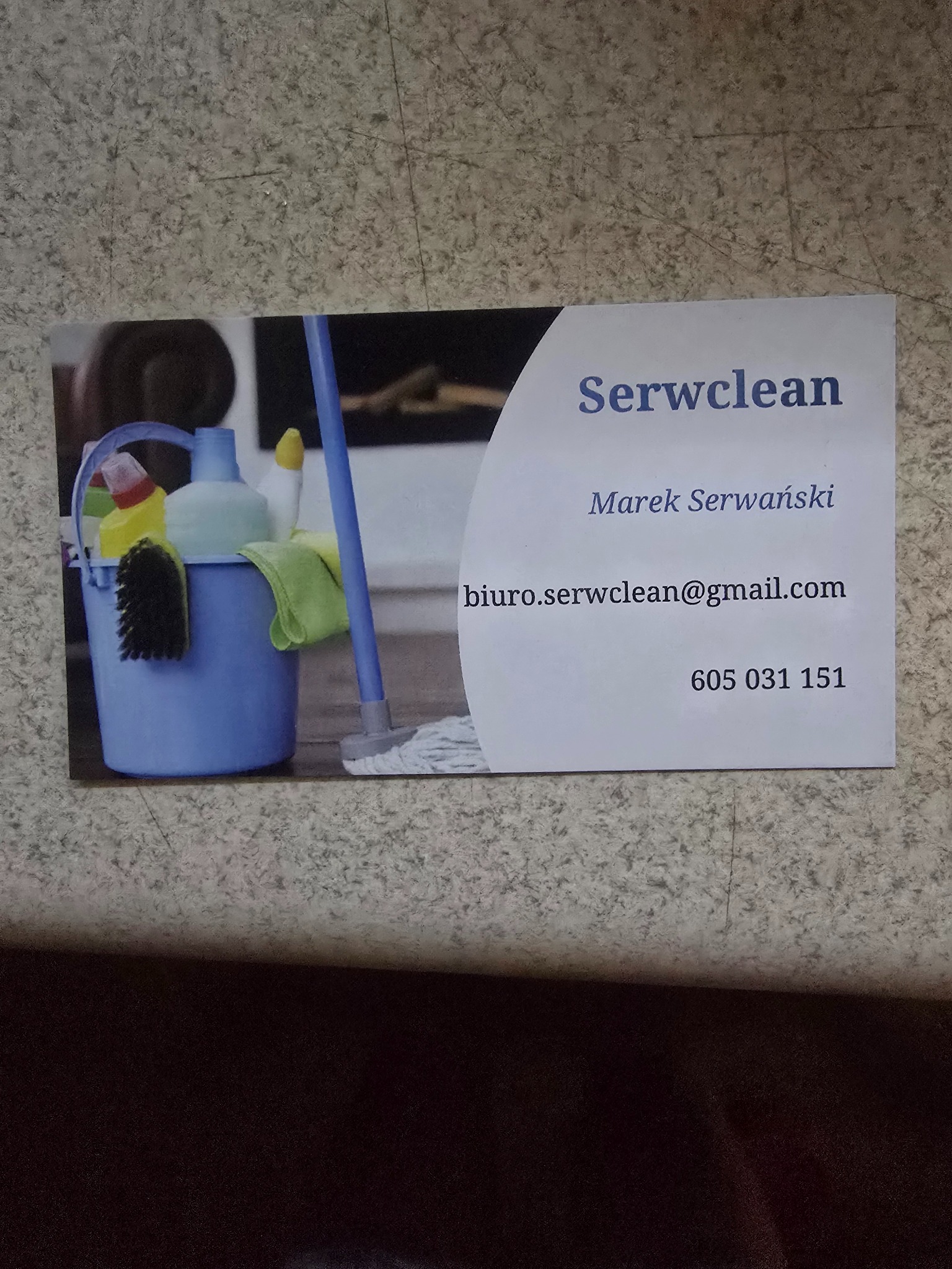 Wizytówka firmy sprzątającej Serwclean z wiadrem, mopem i środkami czystości, leżąca na beżowej powierzchni.