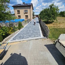 PAN BRUK - Świeżo ułożona, szara kostka brukowa z jasnoszarym obramowaniem tworzy długi podjazd; mężczyzna w słuchawkach zagęszcza powierzchnię mechaniczną ubijarką. W tle budynek z panelami słonecznymi...