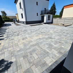 PAN BRUK - Ułożona szara kostka brukowa na podjeździe przed nowoczesnym domem z białymi ścianami i ciemną podmurówką, widoczne studzienki kanalizacyjne i elementy wykończeniowe.