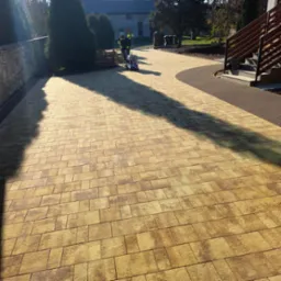 Żółto-brązowa kostka brukowa ułożona na podjeździe, zagęszczana mechanicznie przez pracownika w odblaskowej kamizelce, widoczne palety z kostką i fragment schodów wejściowych.