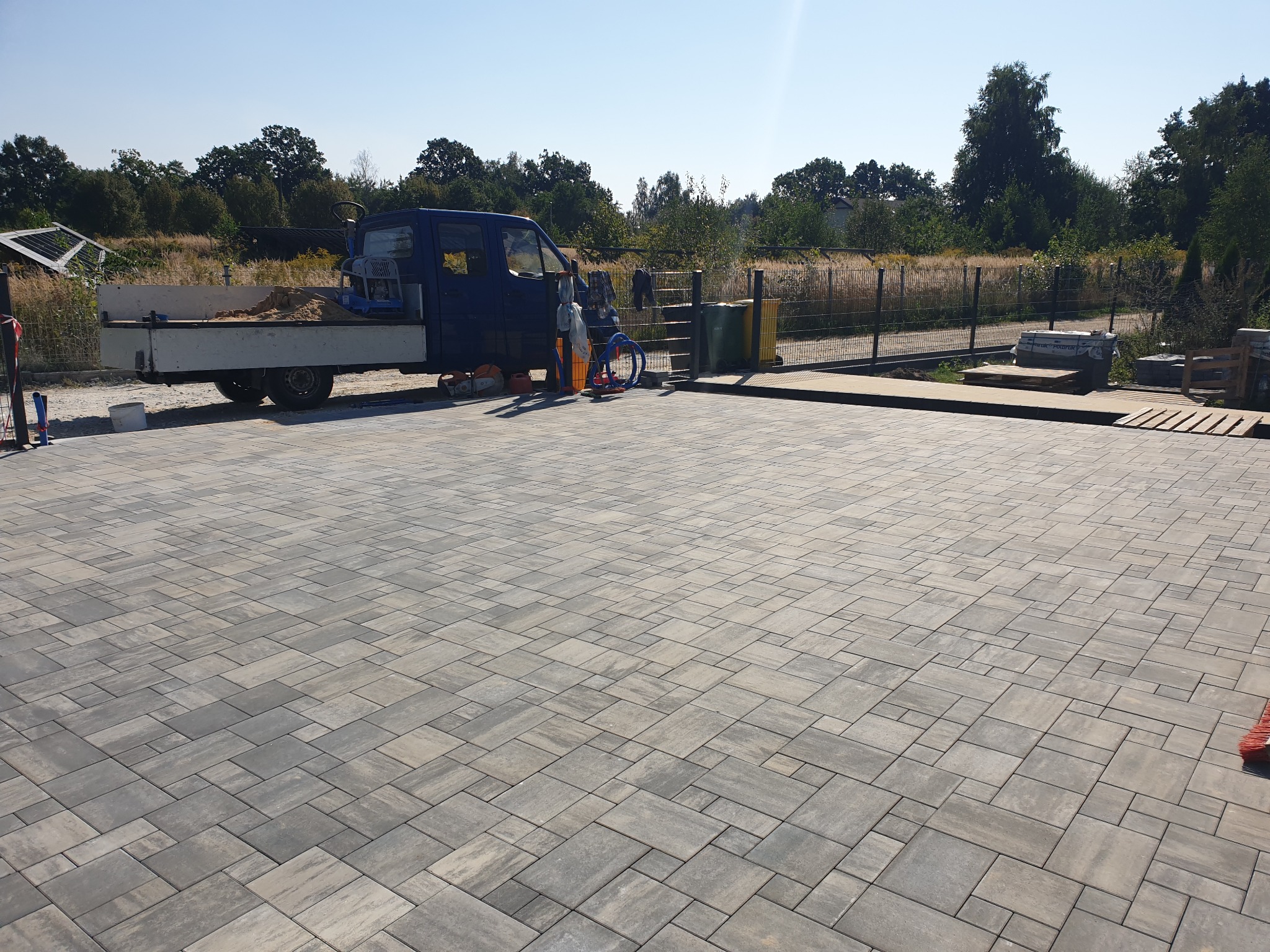 Ukończona powierzchnia wyłożona szarą kostką brukową, widoczny samochód dostawczy z piaskiem, zagęszczarka w tle i narzędzia do pracy, krajobraz z drzewami w oddali.