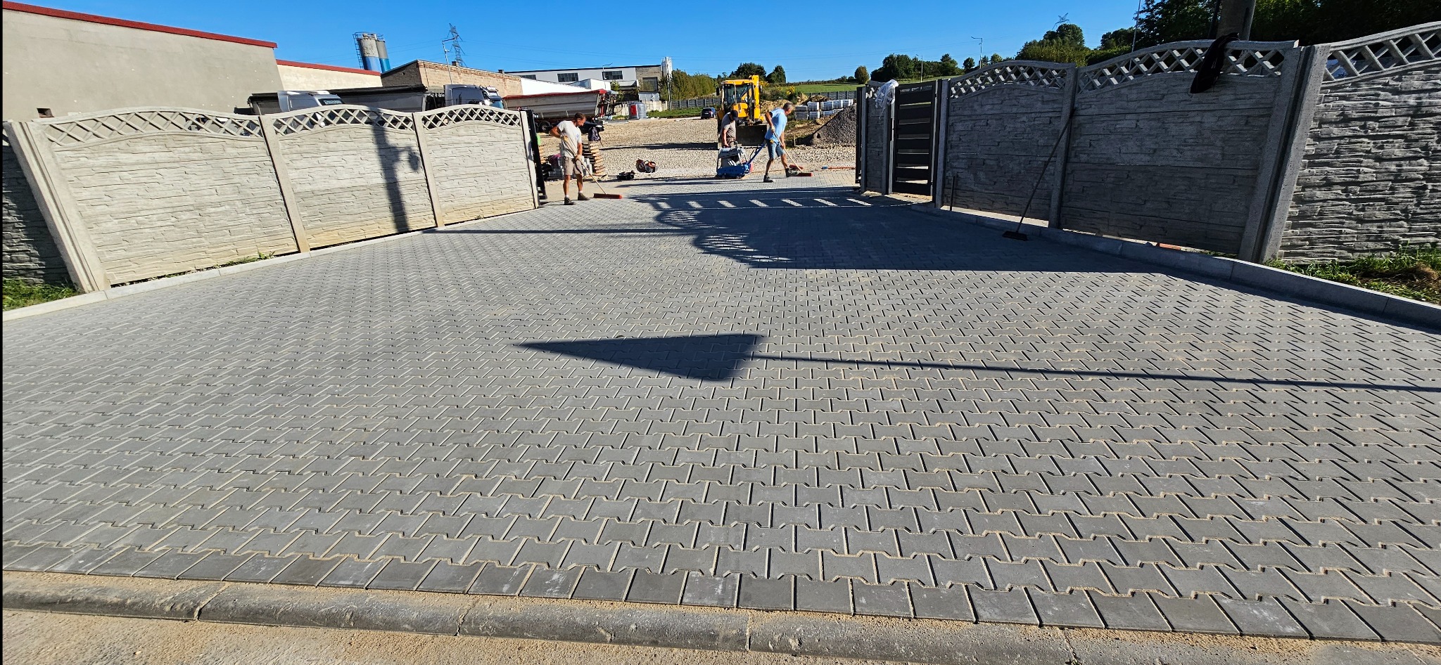 Nowa, szara nawierzchnia z kostki brukowej ułożona na podjeździe, z widocznymi betonowymi panelami ogrodzeniowymi po bokach i pracownikami w tle.
