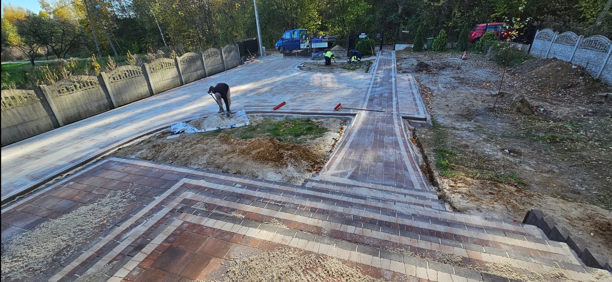 Układanie kostki brukowej na posesji: robotnik wyrównuje piasek pod kostkę, w tle ekipa kontynuuje prace, widoczny częściowo ułożony chodnik i podjazd z kostki w odcieniach brązu i beżu.