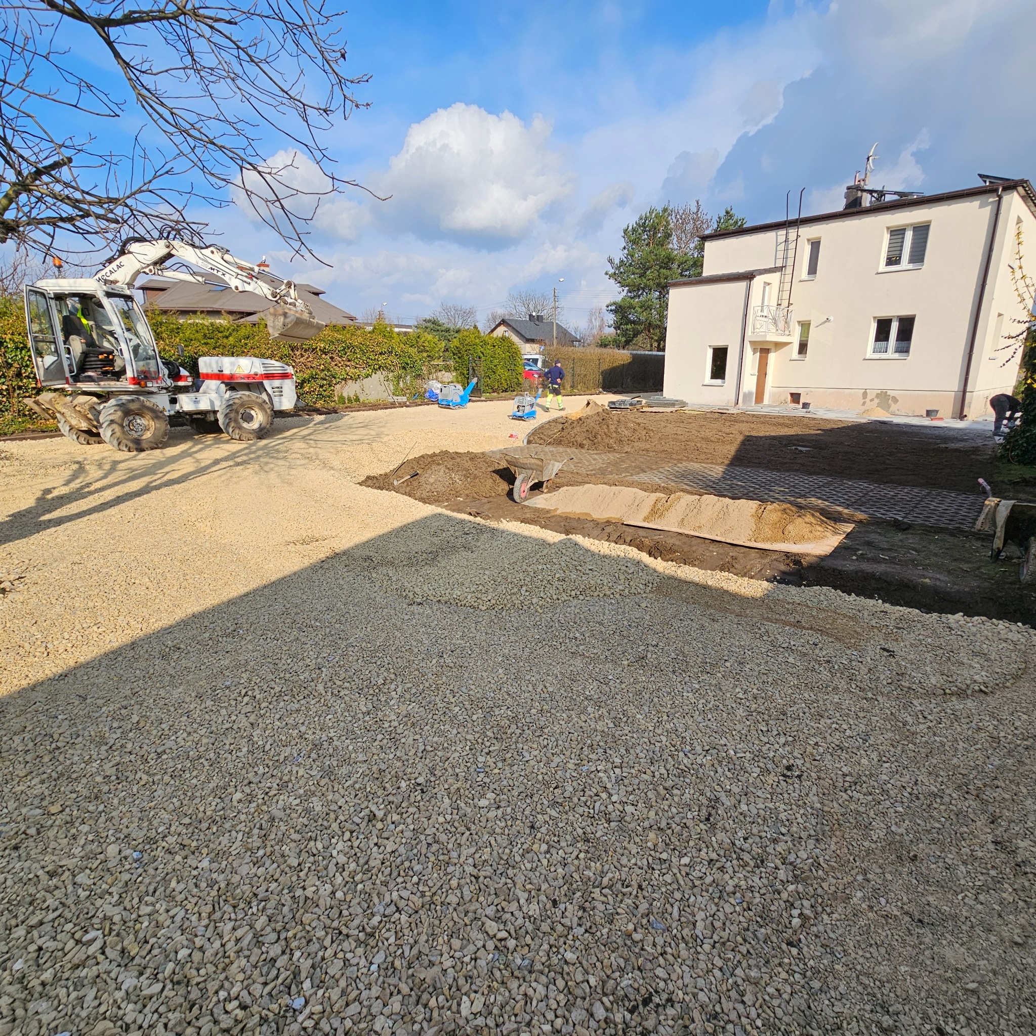 Przygotowanie podłoża pod kostkę brukową z widocznym wykopem, warstwą podsypki żwirowej, częściowo ułożoną kostką ażurową, minikoparką i pracownikami w tle, na tle domu i błękitnego nieba.