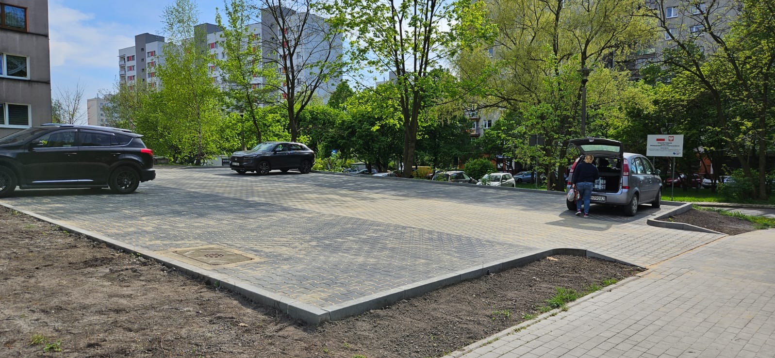 Nowo wyłożony, szary parking z kostki brukowej, z zaparkowanymi samochodami i spacerującą osobą przy otwartym bagażniku, na tle budynków mieszkalnych i zieleni.