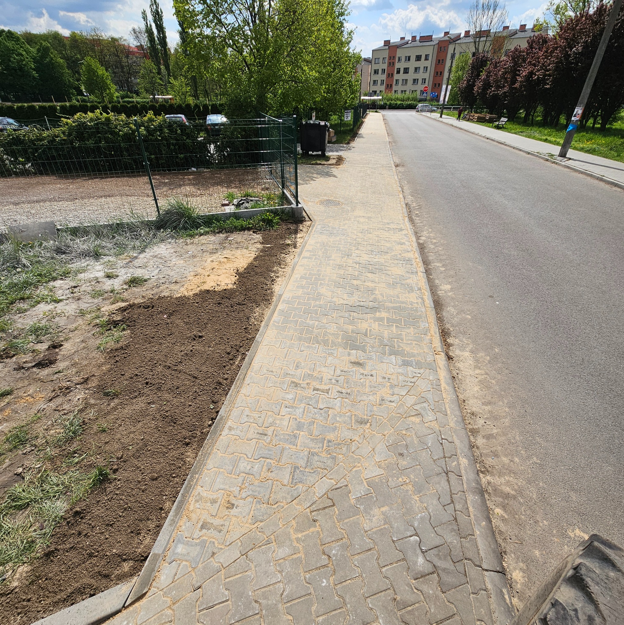 Ułożona kostka brukowa na chodniku, z przygotowanym podłożem z ziemi po lewej stronie, widok wzdłuż chodnika w kierunku ulicy i budynków w tle.
