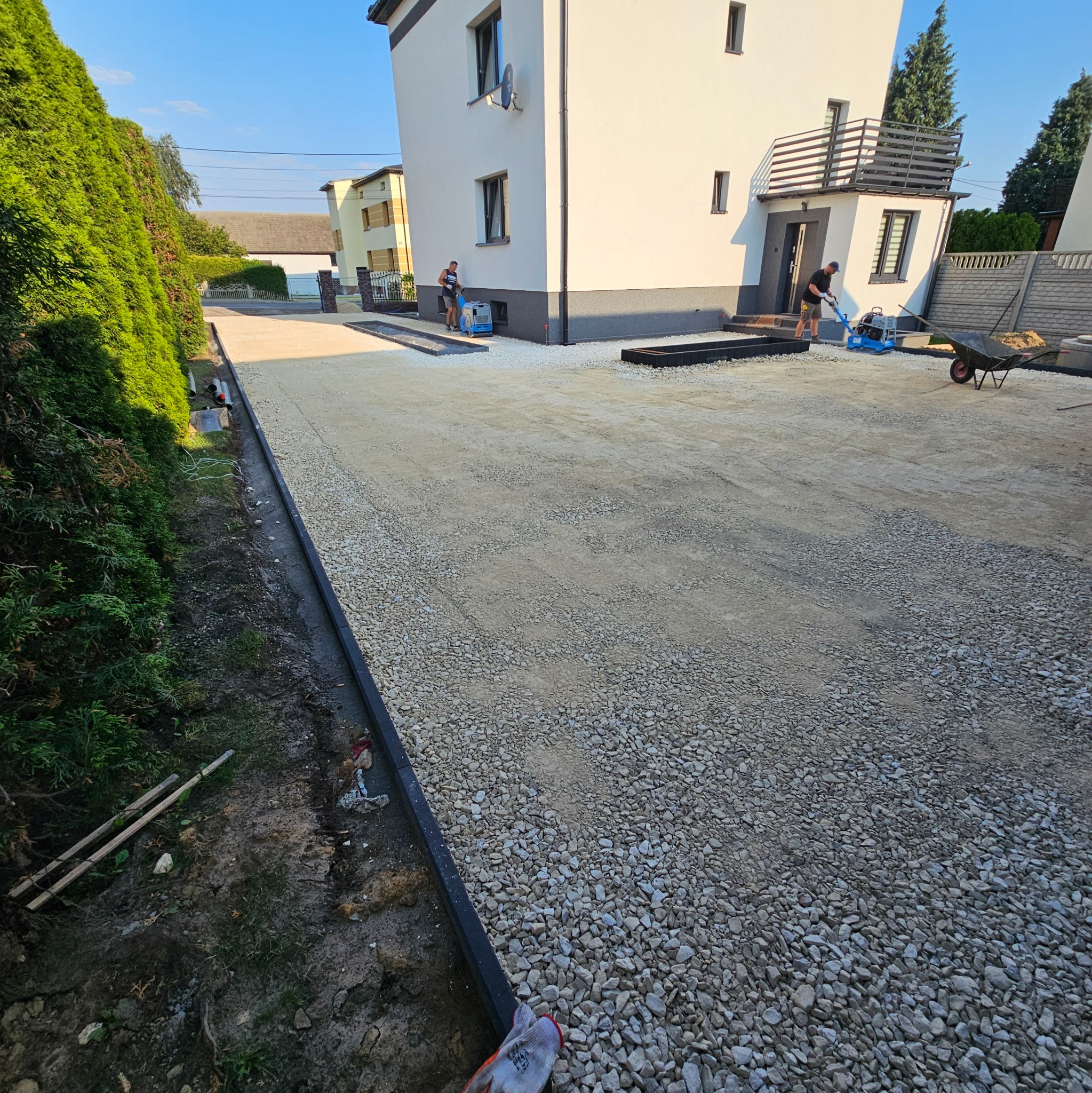 Dwaj mężczyźni pracują na posesji przed domem, jeden ubija tłuczeń zagęszczarką, drugi wyrównuje teren, widoczne obrzeża trawnikowe i przygotowane podłoże pod kostkę brukową.