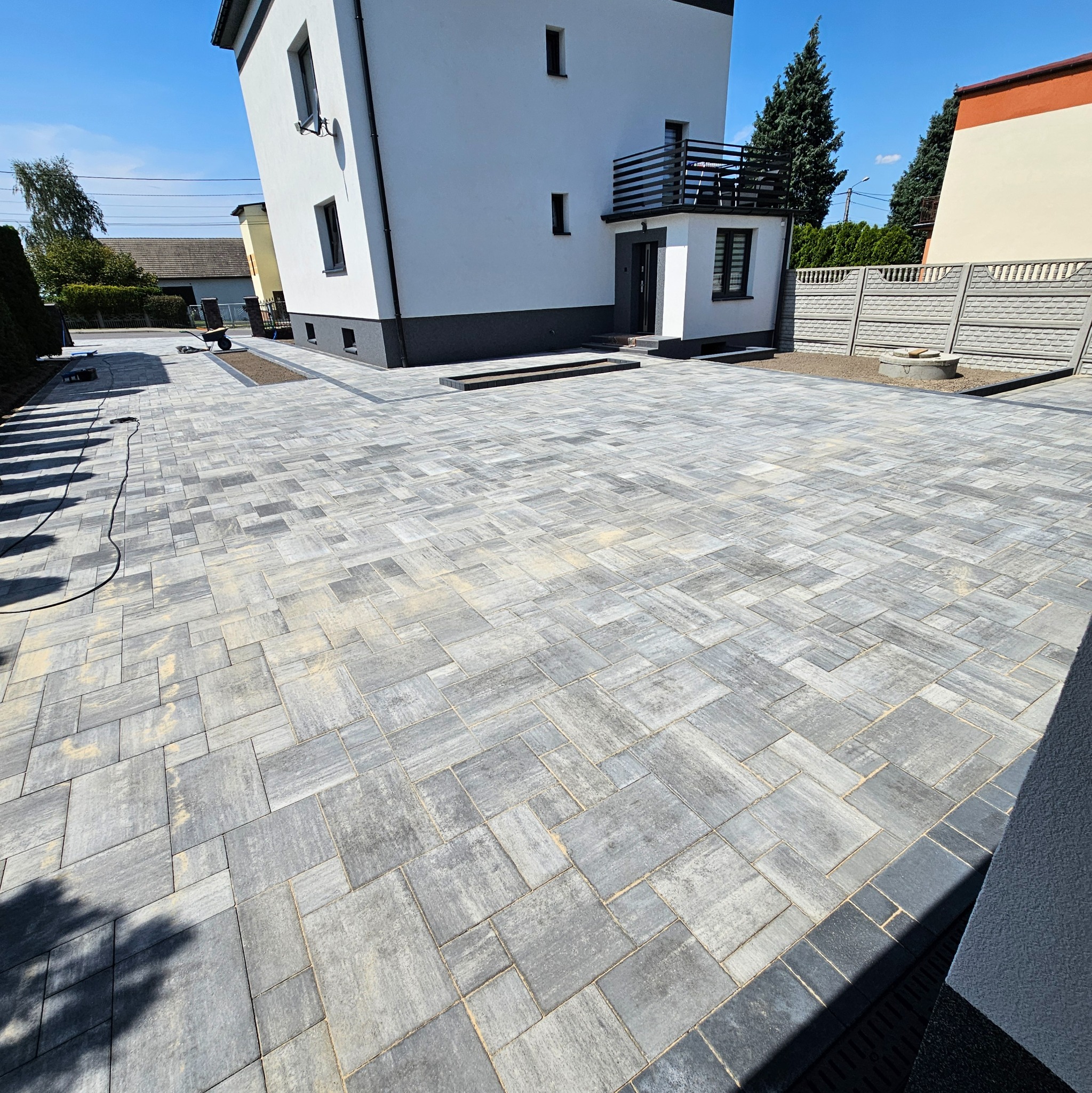 Ułożona szara kostka brukowa na podjeździe przed nowoczesnym domem z białymi ścianami i ciemną podmurówką, widoczne studzienki kanalizacyjne i elementy wykończeniowe.