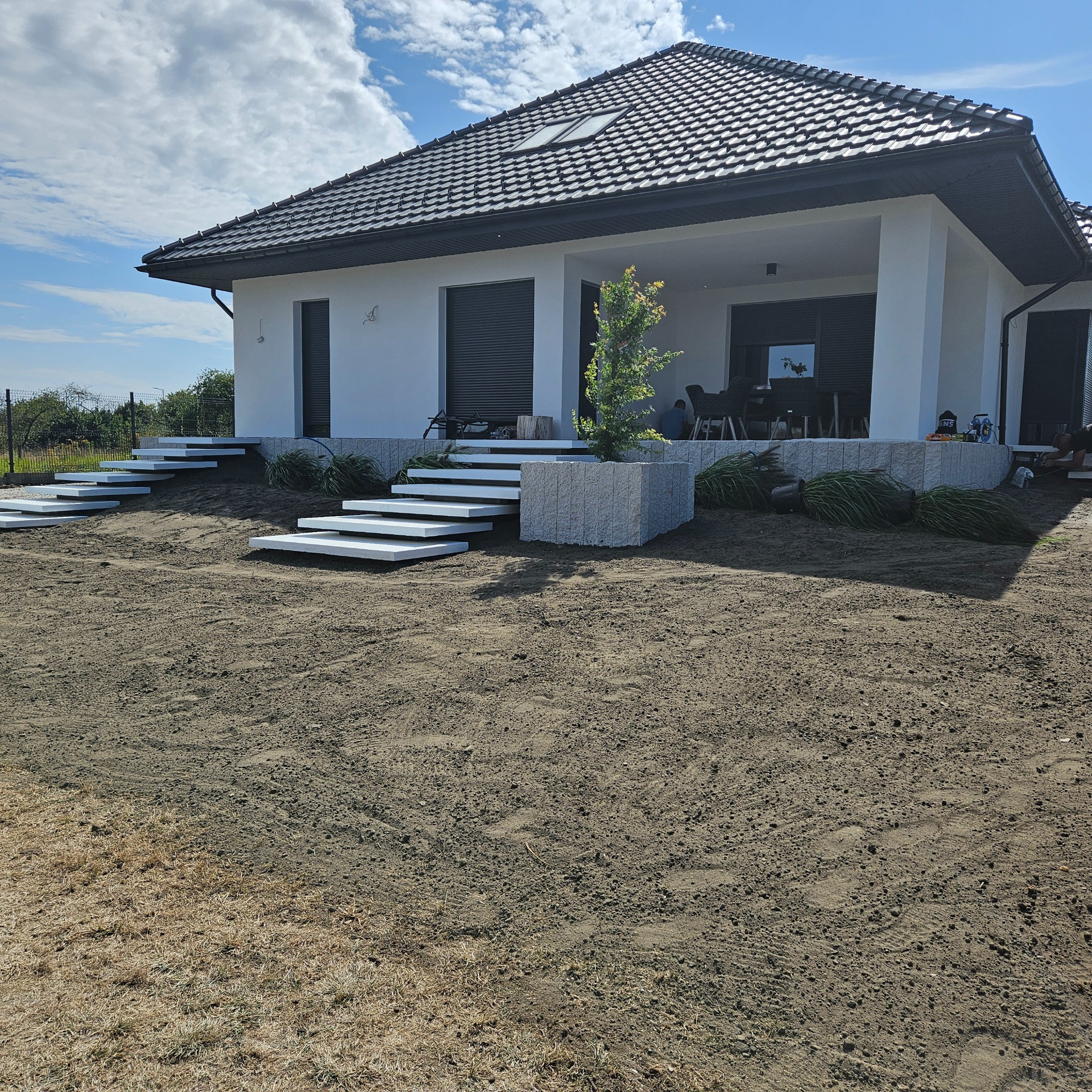 Nowoczesny dom jednorodzinny z białymi schodami prowadzącymi do wejścia, teren wokół wyrównany i przygotowany pod trawnik, widoczne donice z roślinami i osoba pracująca przy wykończeniu.