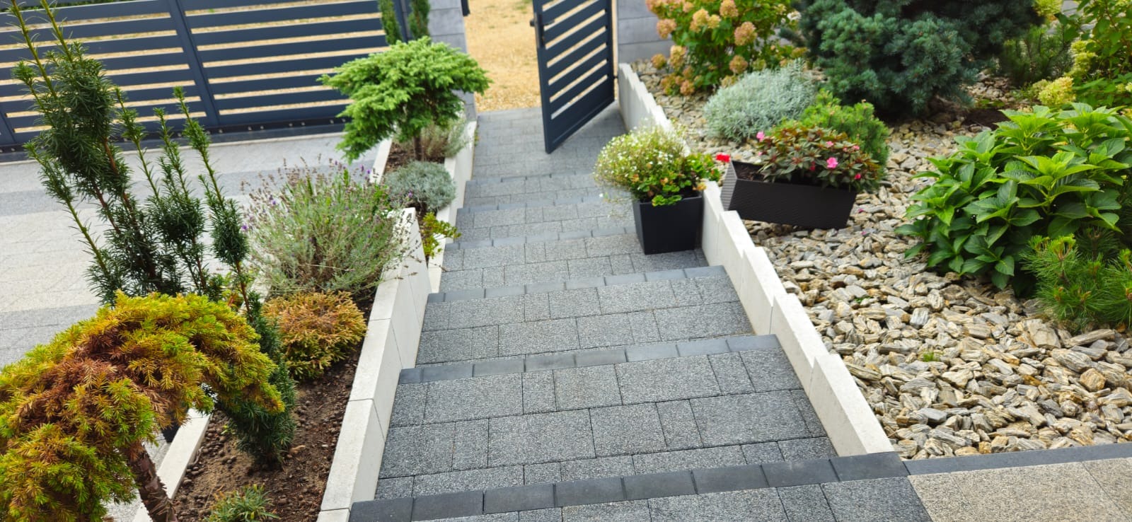 Schody z szarej kostki brukowej prowadzące do furtki, otoczone roślinnością ozdobną i wysypane kamieniami, z widocznymi donicami z kwiatami po bokach.