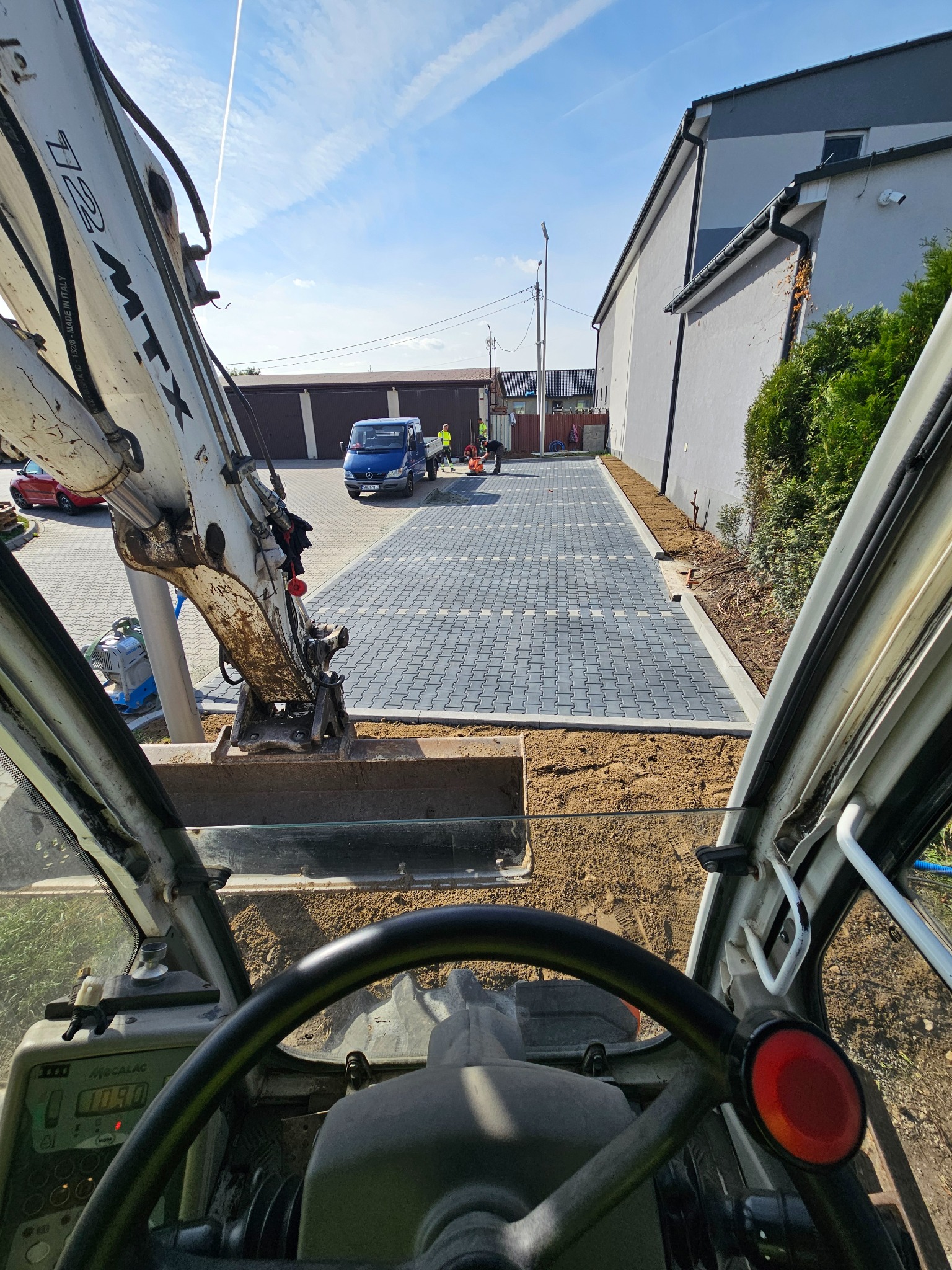 Widok z kabiny koparki na świeżo ułożoną, szarą kostkę brukową na podjeździe, z widocznymi pracownikami i niebieskim samochodem dostawczym w tle, w słoneczny dzień.