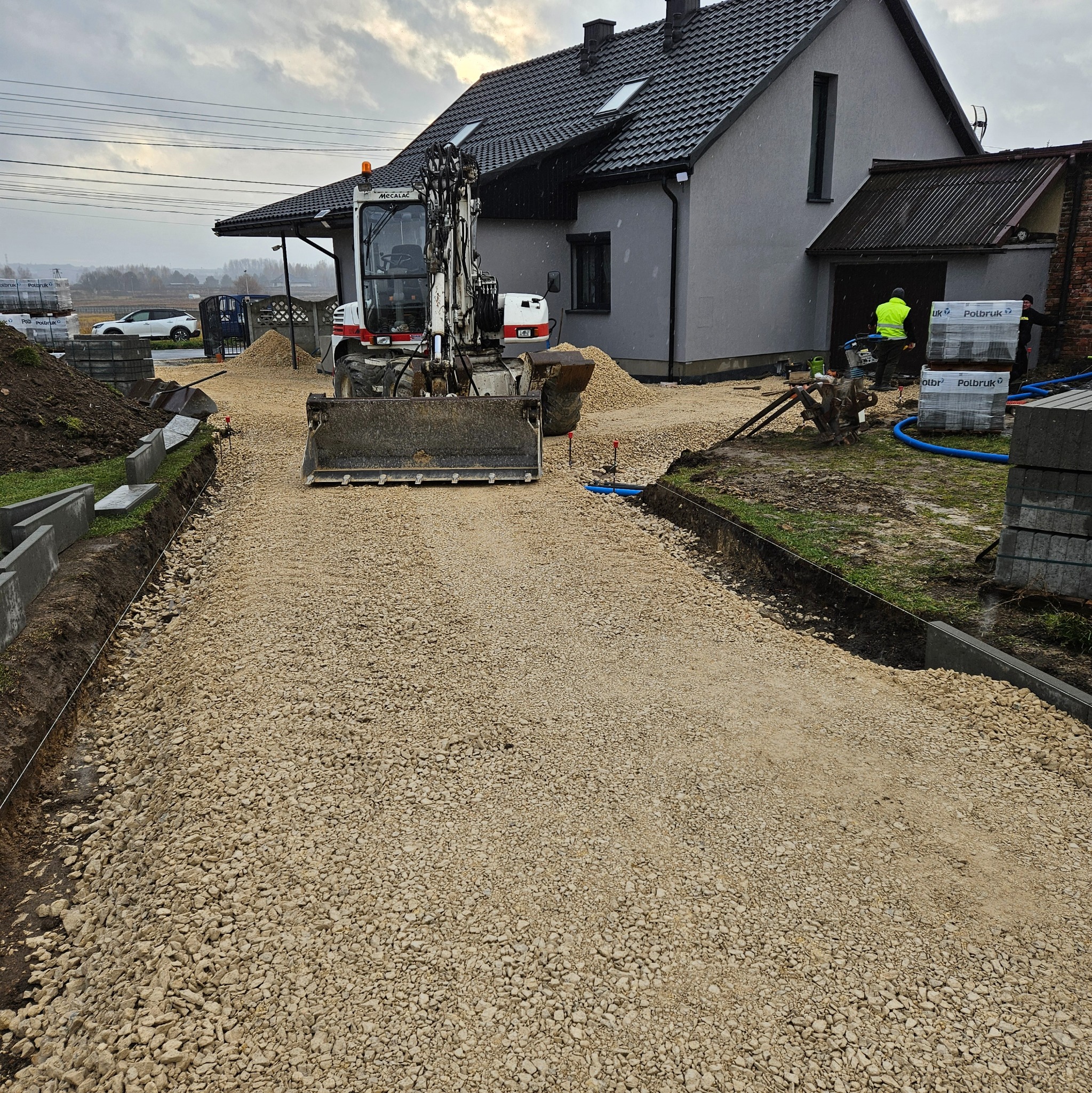 Przygotowany podjazd z wysypanym kruszywem, oczekujący na dalsze prace, z widoczną koparką Mecalac, paletami kostki Polbruk i pracownikami w tle.