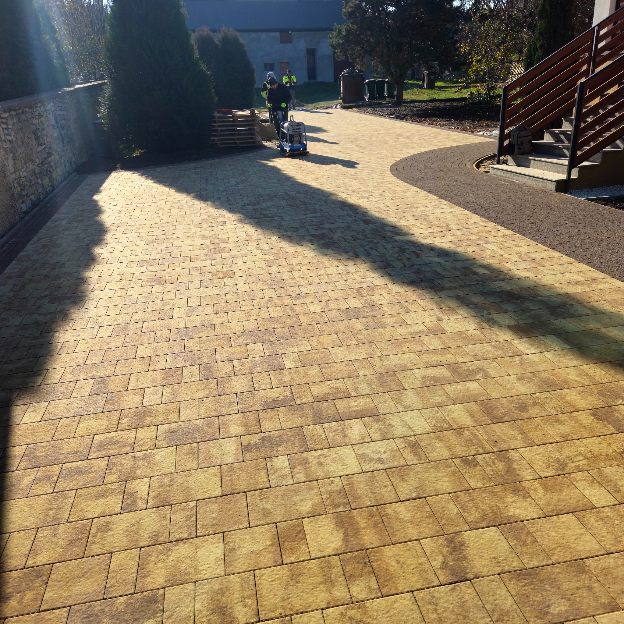 Żółto-brązowa kostka brukowa ułożona na podjeździe, zagęszczana mechanicznie przez pracownika w odblaskowej kamizelce, widoczne palety z kostką i fragment schodów wejściowych.