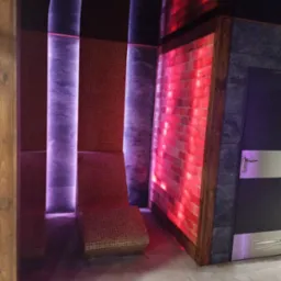 Nowoczesne wnętrze sauny z mozaikową ławką, ścianą solną w drewnianej ramie i podświetleniem LED w odcieniach fioletu i czerwieni, obok drzwi z metalowymi akcentami.