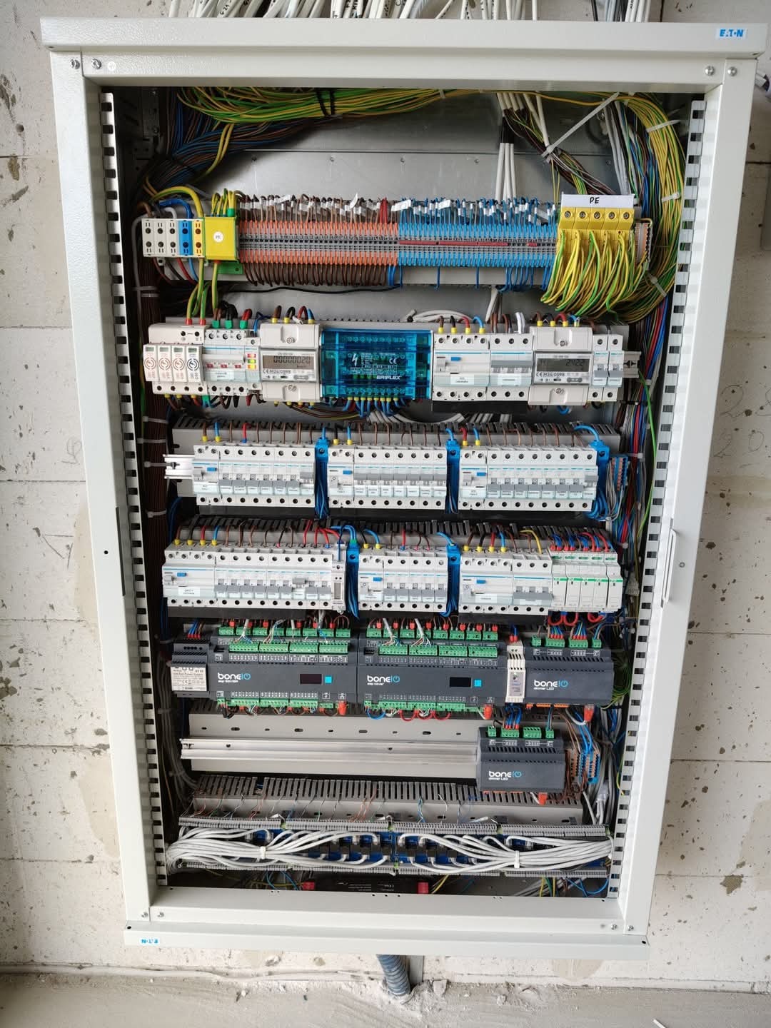 Rozdzielnica elektryczna w metalowej obudowie, widoczne liczne kable, bezpieczniki i moduły. Instalacja schludna, uporządkowana, zamontowana na szarej ścianie.