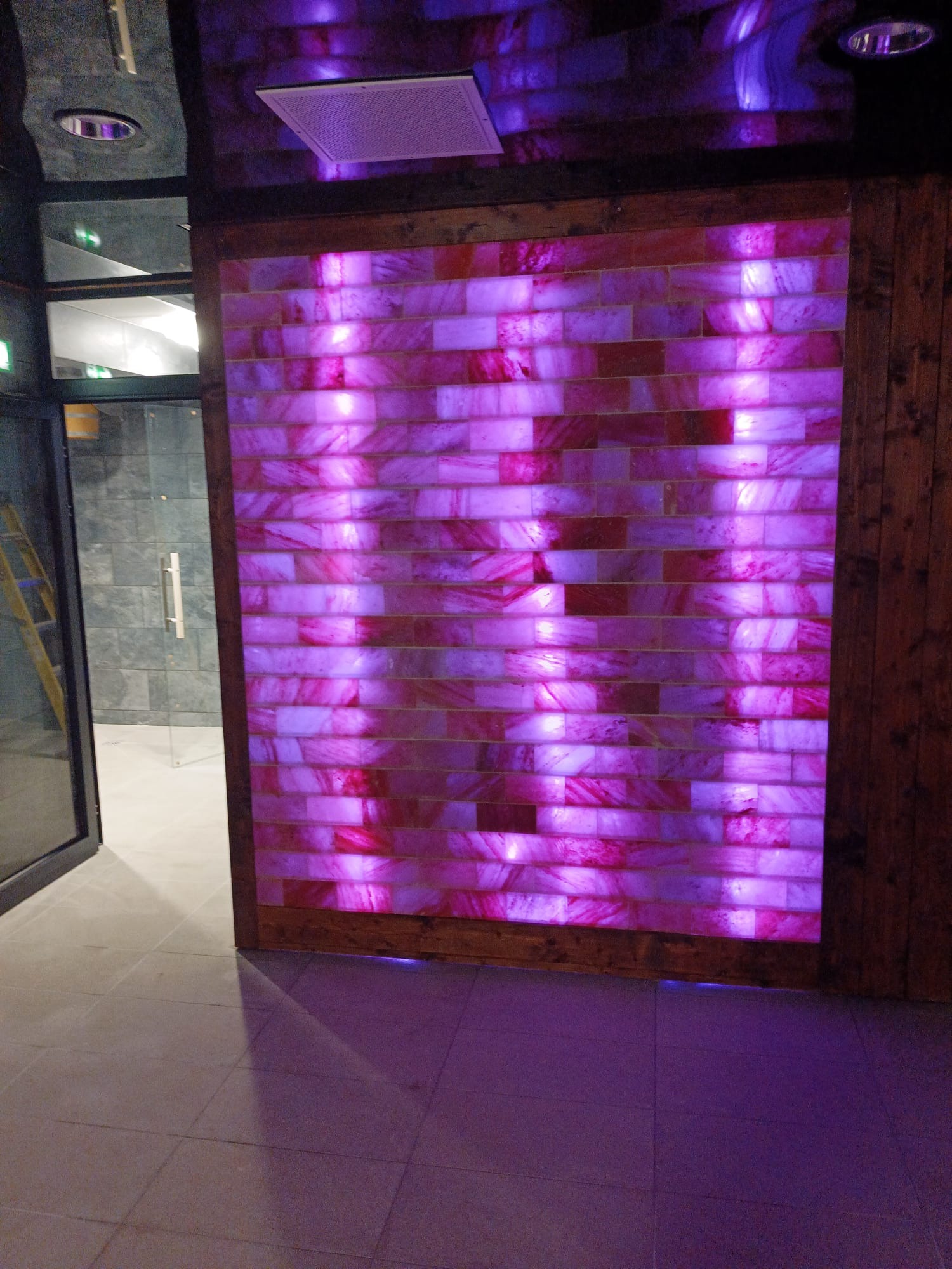 Podświetlana, różowa ściana z bloków soli himalajskiej w drewnianej ramie, widoczna z perspektywy wnętrza pomieszczenia z drzwiami i schodami w tle.