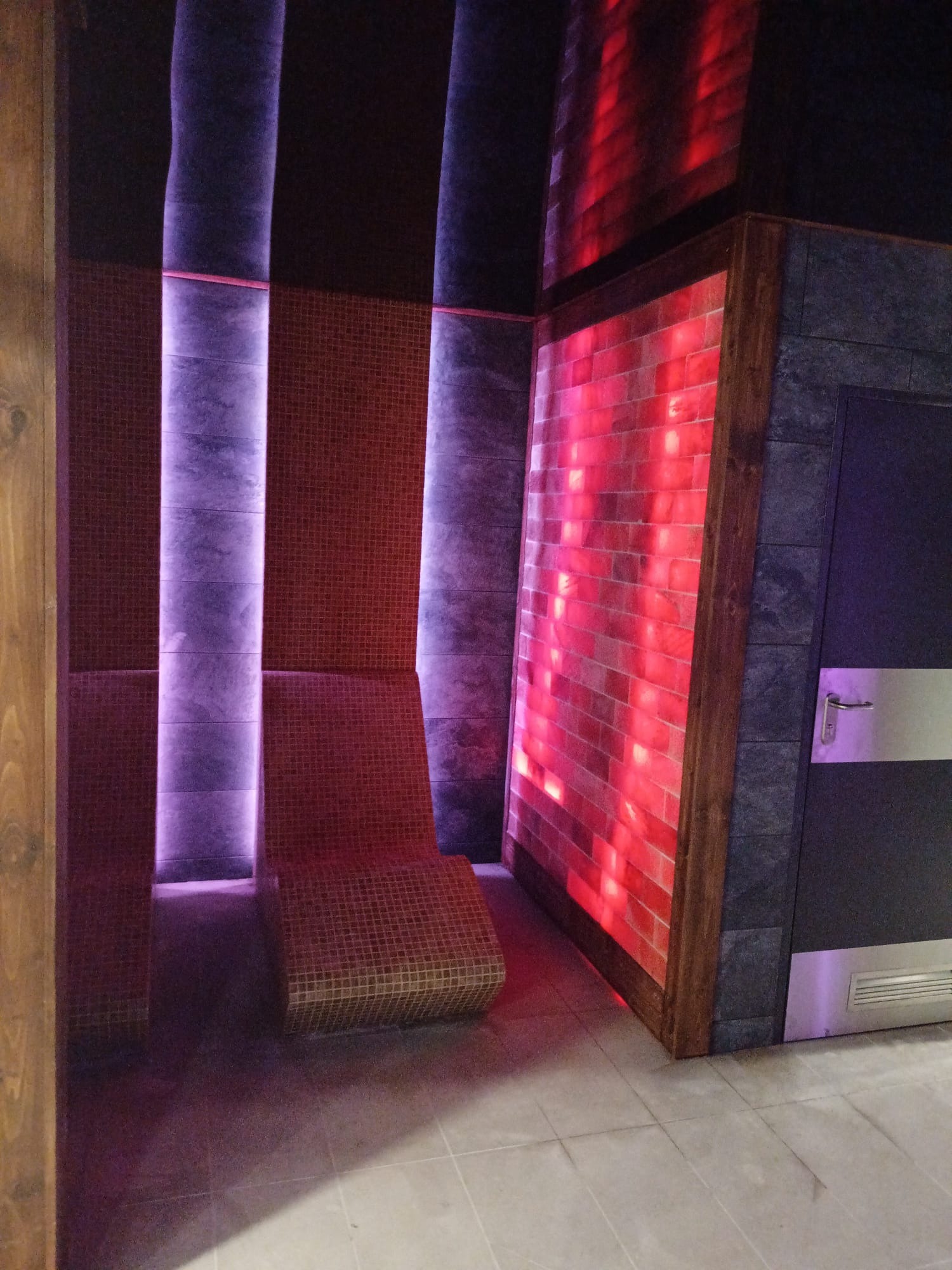 Nowoczesne wnętrze sauny z mozaikową ławką, ścianą solną w drewnianej ramie i podświetleniem LED w odcieniach fioletu i czerwieni, obok drzwi z metalowymi akcentami.