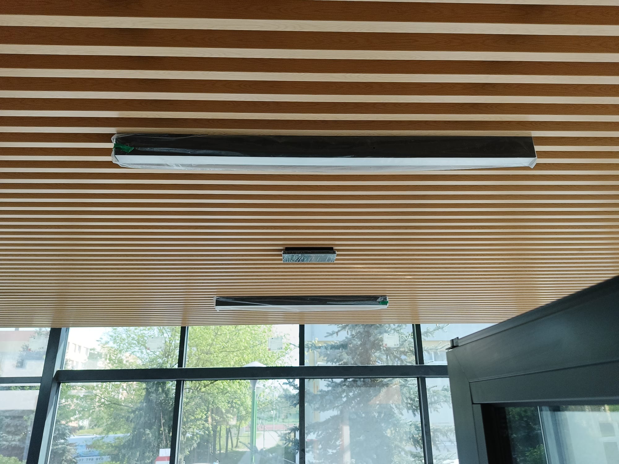 Montaż opraw oświetleniowych LED na drewnianym suficie lamelowym w nowoczesnym biurze, widok z dołu na okna z widokiem na zieleń.