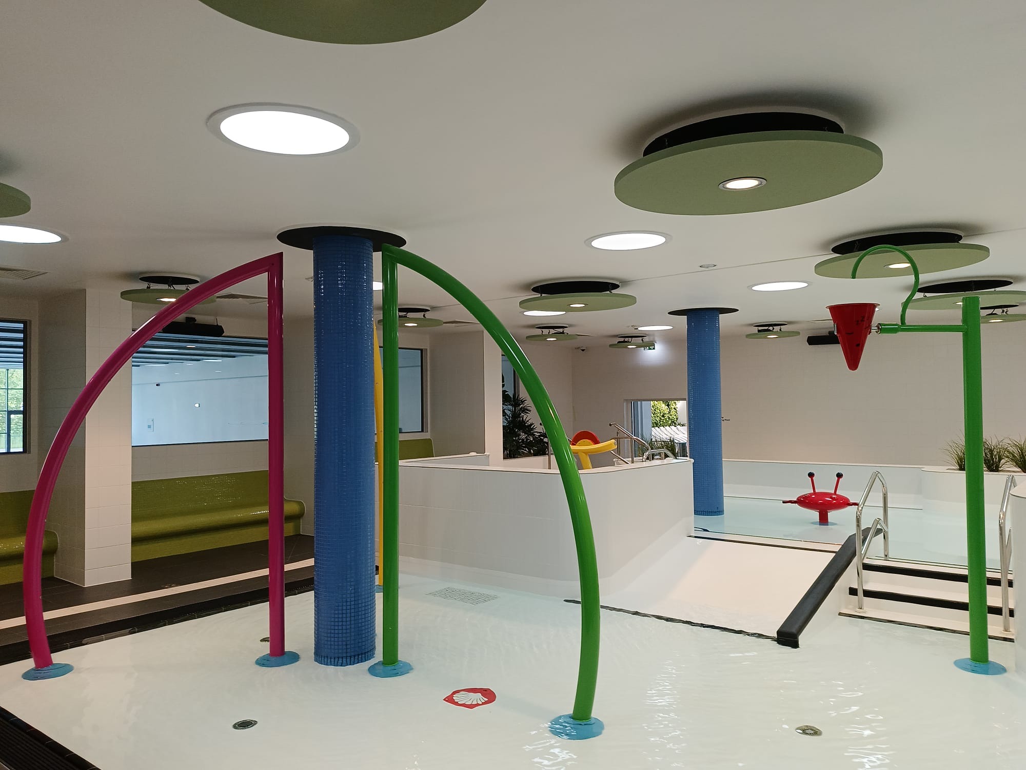 Nowoczesny basen rekreacyjny z kolorowymi elementami i oświetleniem punktowym. W tle widoczne okna i minimalistyczne wykończenie wnętrza.