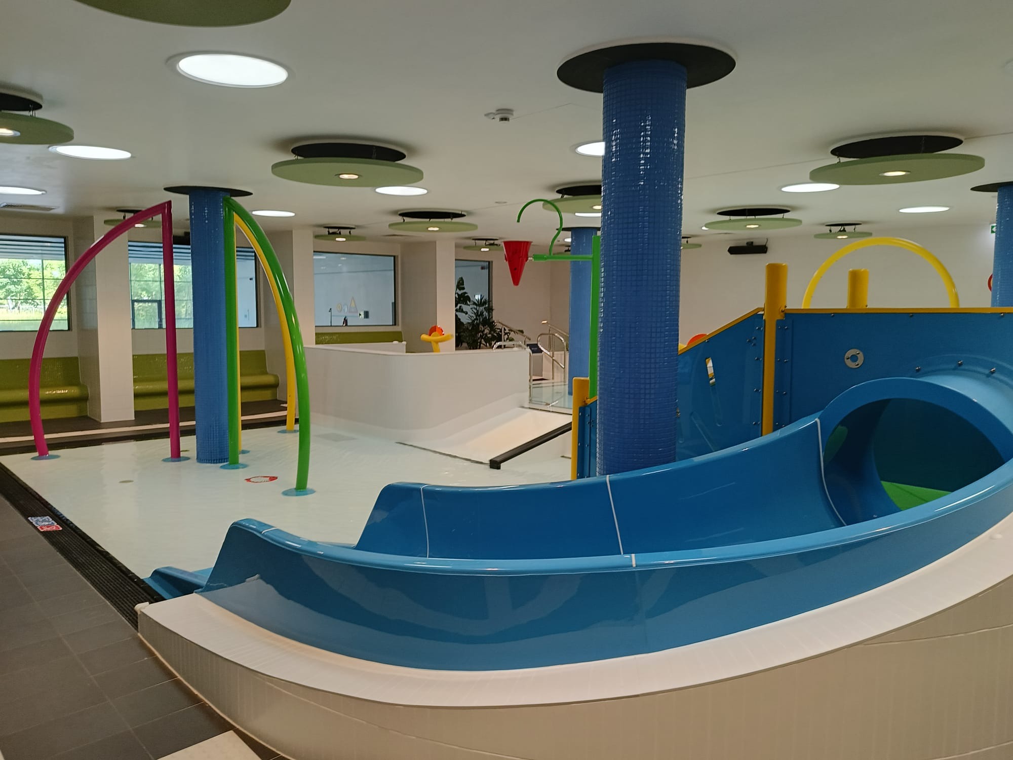 Kryty basen z niebieską zjeżdżalnią, kolorowymi elementami dekoracyjnymi i oświetleniem sufitowym. Widok na płytki basenowe i część wypoczynkową.