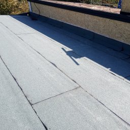 Vasil Irhovtsii - Szary, płaski dach pokryty papą termozgrzewalną z widocznymi łączeniami. W tle fragment ściany z płyt OSB i cień osoby.