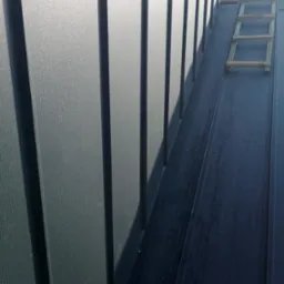 Szary dach z blachy trapezowej w pionowym ujęciu z drewnianą drabiną. Widoczne mocowania blachy. Dach oświetlony słońcem.