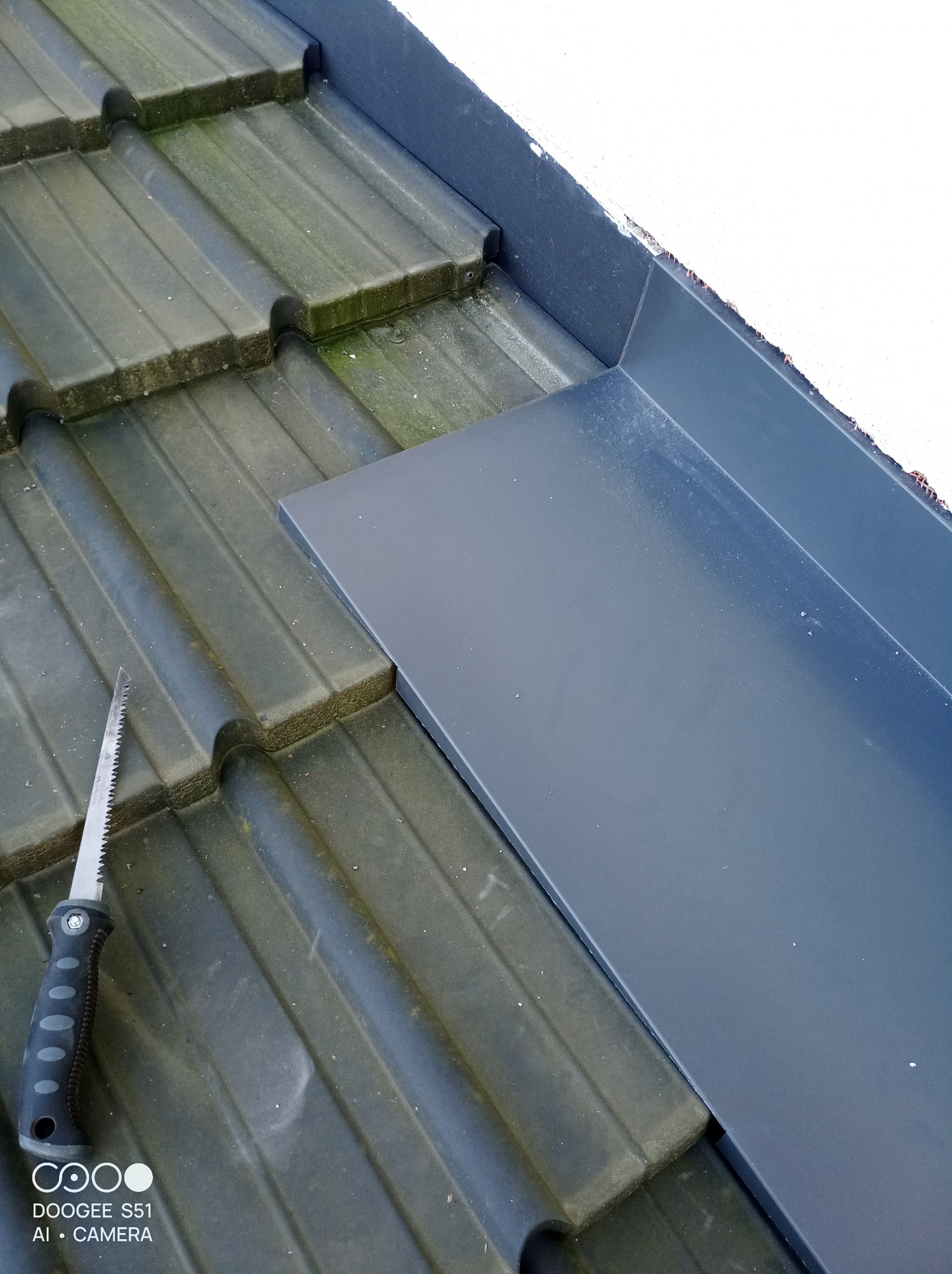 Szara obróbka blacharska przy kominie na tle starej dachówki. Obok leży piła ręczna. Detal z bliska.